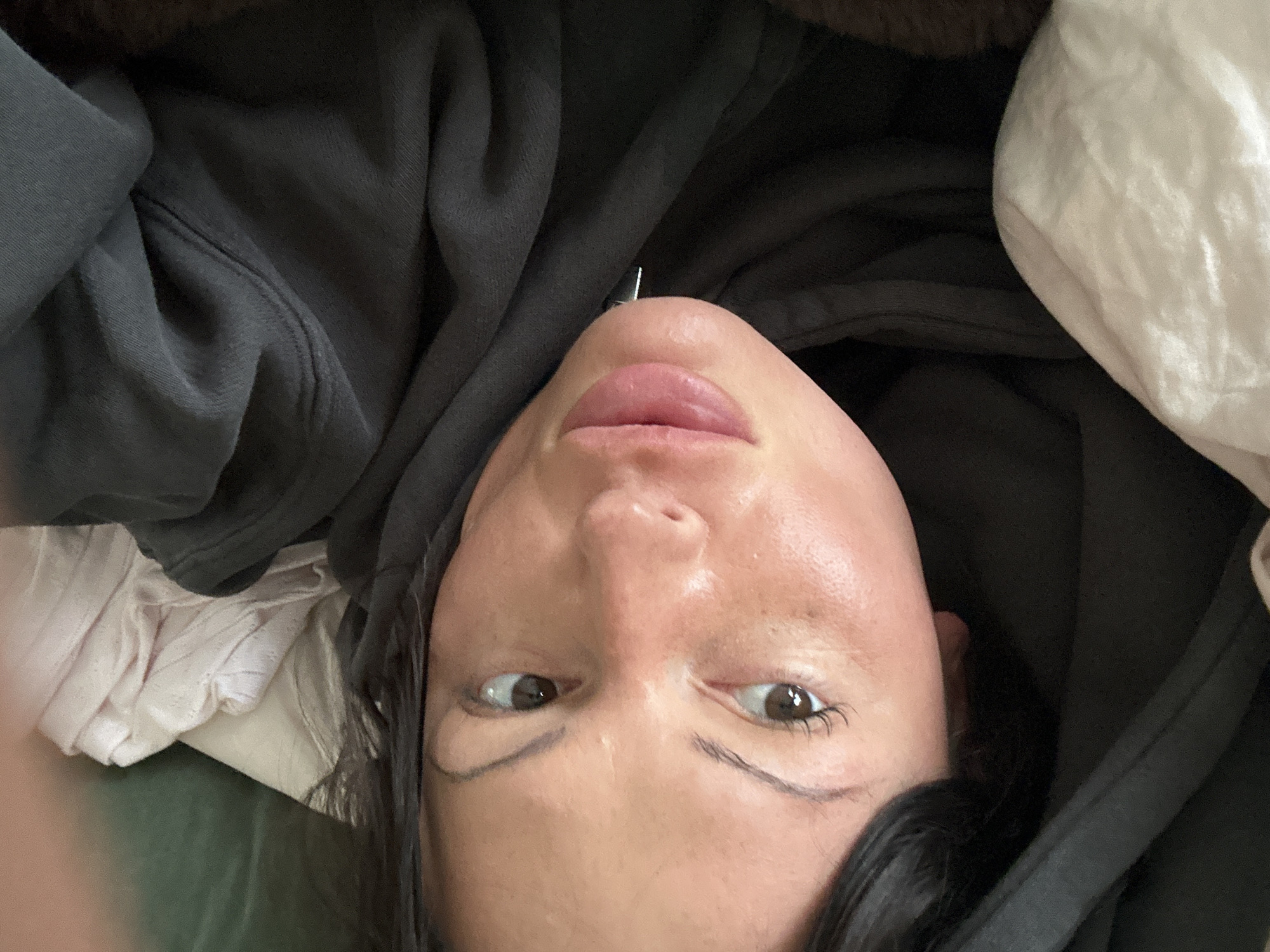 A selfie photograph shows a person lying down, angled towards the camera. The person has fair skin, brown eyes, dark hair, and full lips. The person is wearing a dark gray hooded sweatshirt. A white pillow is visible to the right of the person's head. The location of Tunbridge Wells, United Kingdom cannot be confirmed from the image.
