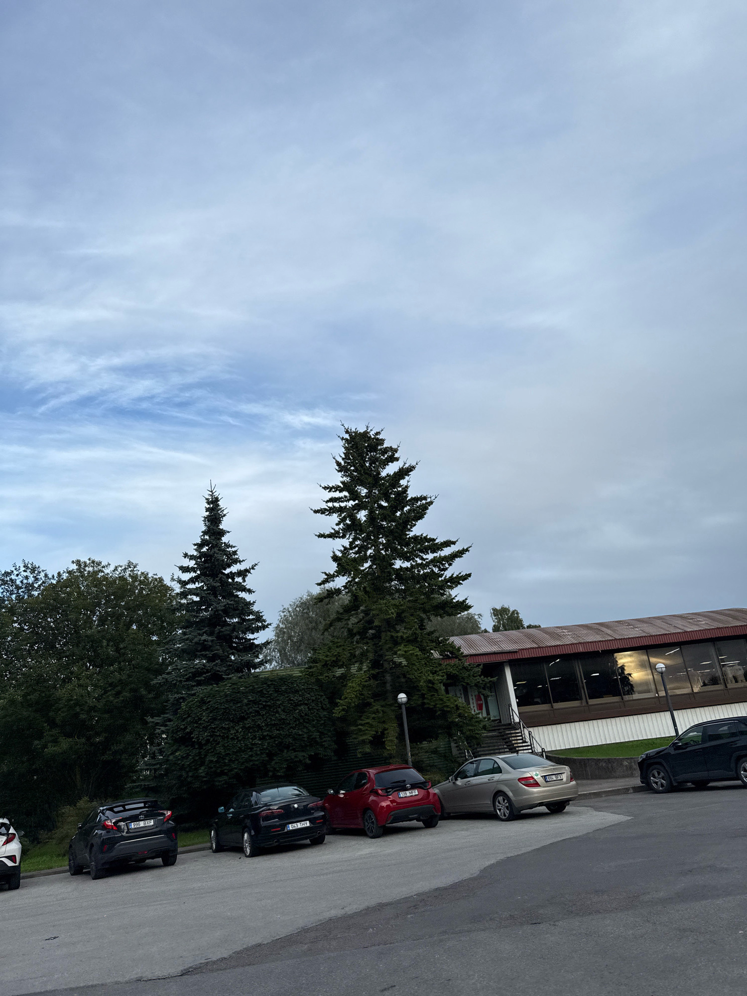 The image shows a parking lot with several cars parked in front of a building. The cars are of various models and colors, including a black Toyota, a black Alfa Romeo, a red Toyota, a gold Mercedes-Benz, and a dark SUV. There are also several trees and bushes in the background, as well as a building with large windows. The sky is overcast with some clouds. Based on the license plates, the image appears to have been taken in Estonia. Specifically, the license plates "996 BXP", "043 THY", "138 MPP", and "ESL BF1" are consistent with Estonian license plates. The building in the background resembles a typical structure found in industrial or commercial areas of Tallinn, Estonia.