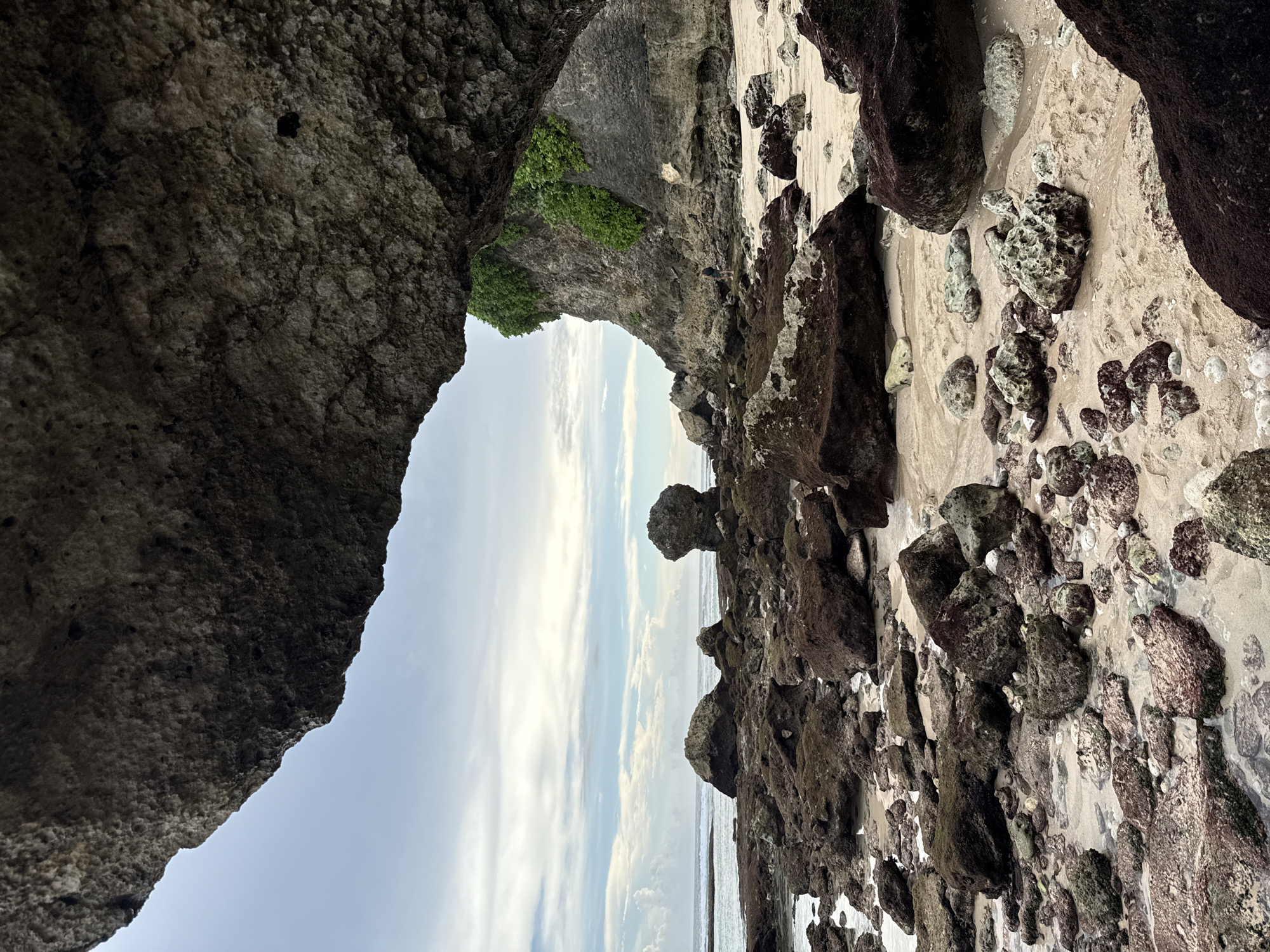 A coastal scene in Ungasan, Indonesia. The image is composed from inside a rock arch looking outward to the ocean, with the sky visible through the arch. The foreground is a sandy beach with a mixture of large dark rocks and smaller stones scattered across it. The sand appears light in color, contrasting with the darker rocks. The rock arch framing the view is gray and rough-textured. Some green vegetation is visible on the rock face within the arch. In the background, the ocean meets the sky, creating a horizon line. The sky contains some clouds. The image is taken from a low angle. There are no people visible in the image.