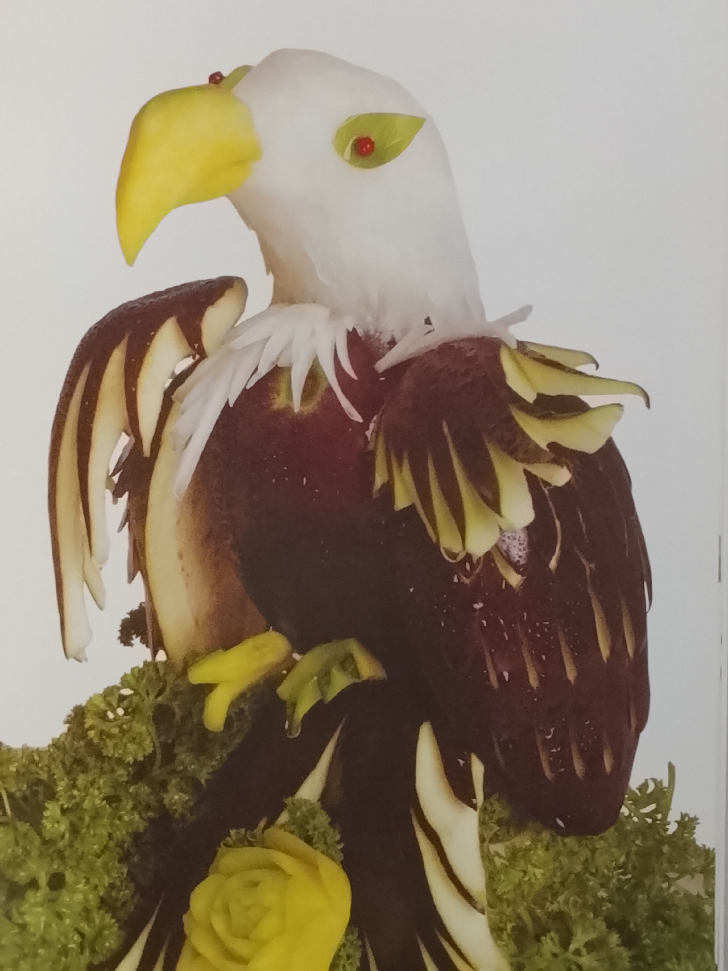 A close-up shot showcases a vegetable carving of an eagle. The eagle's head and neck are white, the beak is yellow, and the eyes are marked with small red spots. Its body is brown, and the wings are comprised of dark brown vegetable slices with alternating light-colored strips. White vegetable strips resembling feathers are positioned around its neck. The eagle stands on a bed of green leafy vegetables resembling shrubbery. A yellow vegetable carving in the shape of a rose is visible near the base of the vegetable eagle. The background is plain white. The image shows no evidence of Altagracia, Venezuela.