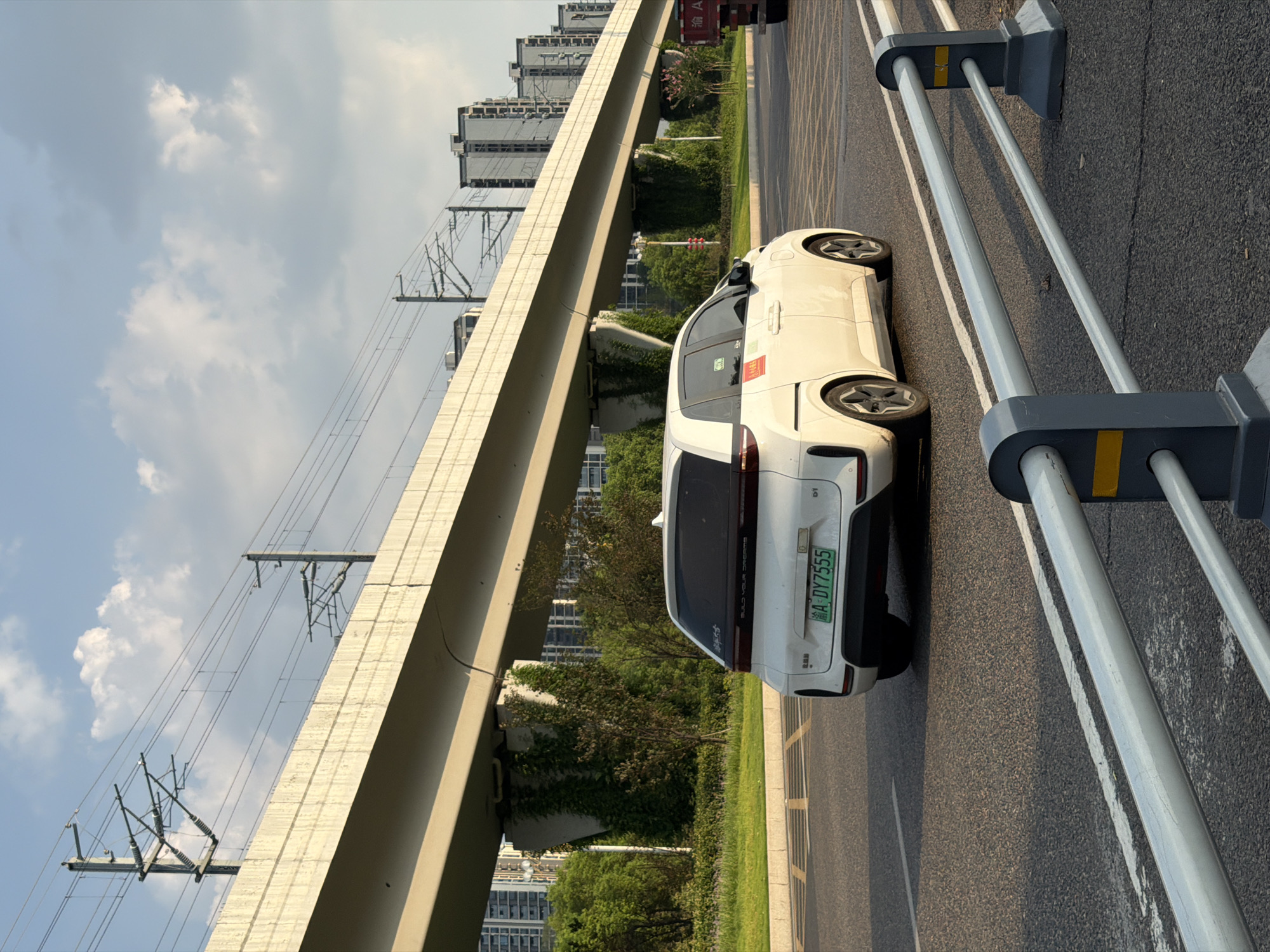 A white electric vehicle is driving on a paved road under a concrete overpass with overhead electrical wires. The vehicle, with license plate "A DY7555", has red and white stickers on its rear door. Greenery and trees line the side of the road, and several tall buildings are visible in the background. The sky is partly cloudy. This scene appears to be in the city of Jiangjin, China.