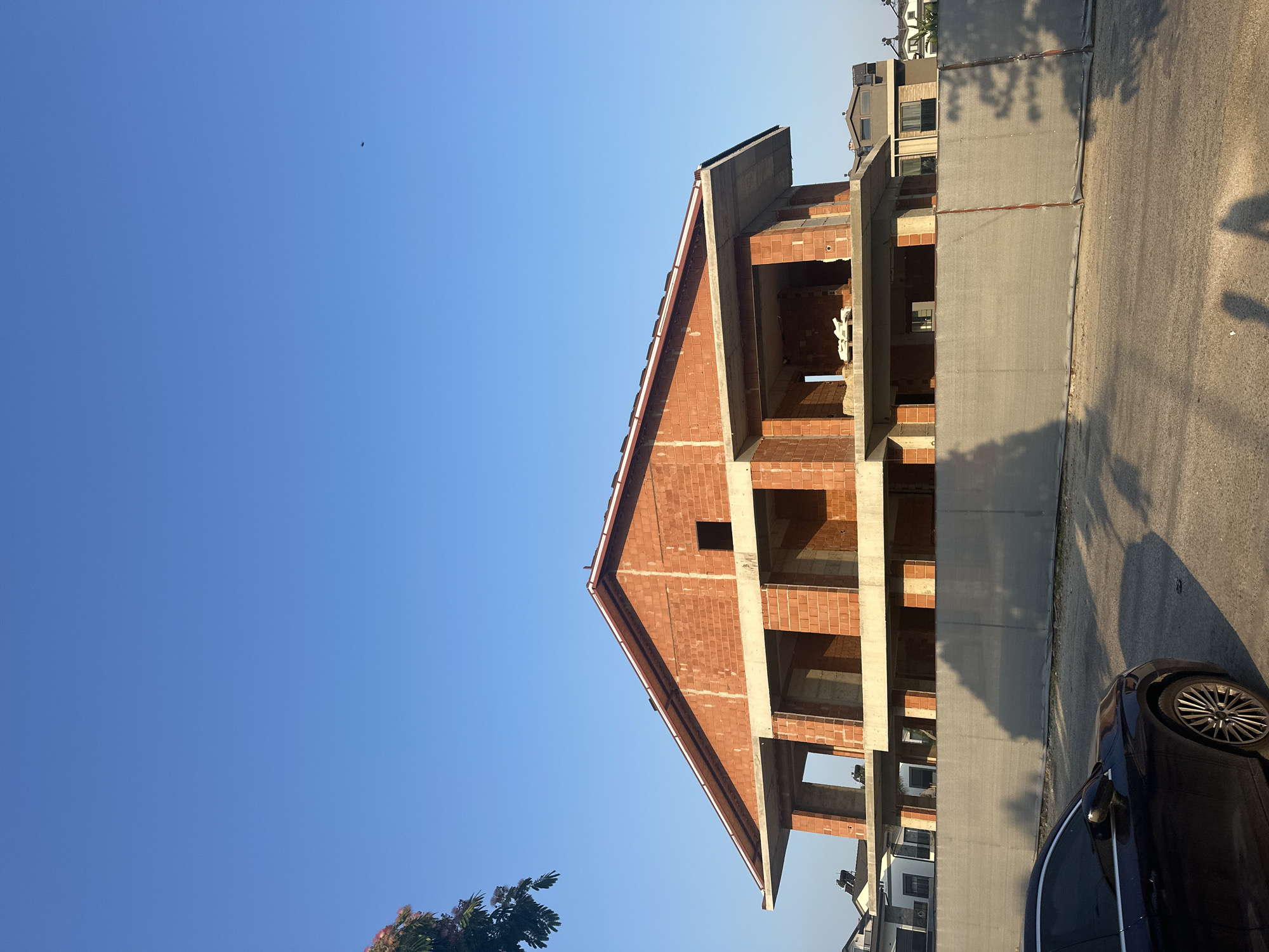 A construction site in the city of Fethiye, Turkey, displays an unfinished brick house with open doorways and windows under a clear blue sky. A dark-colored sedan is parked in the foreground, casting a shadow on the paved surface. A grey construction fence runs across the middle of the image, separating the road from the building site. Nearby, a tree with pink blossoms adds a touch of color to the scene. The surrounding area shows other residential buildings, suggesting a suburban or developing neighborhood. The lighting indicates it is either early morning or late afternoon, with the sun casting long shadows.