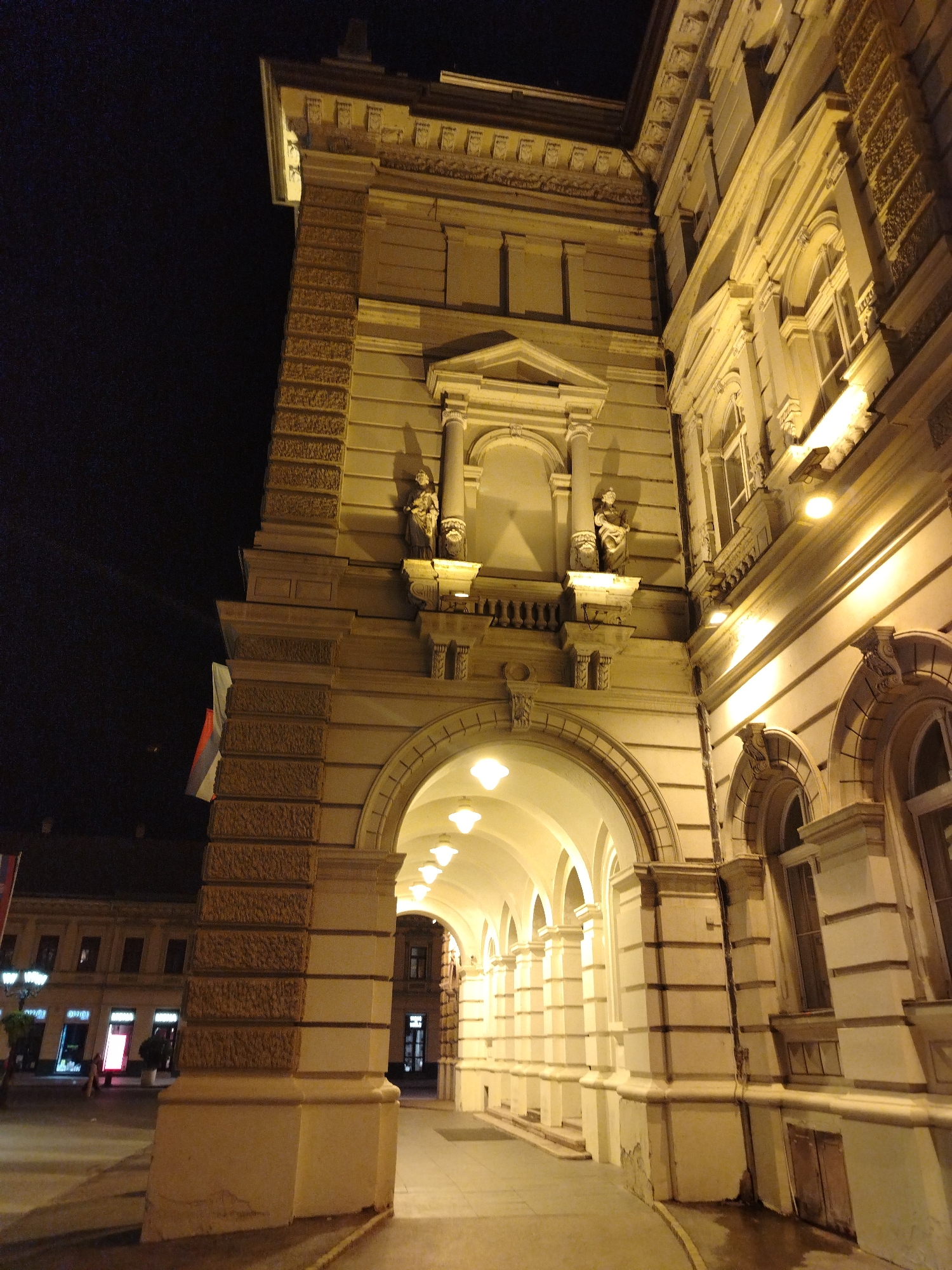 The image depicts the facade of a grand building in Novi Sad, Serbia, at night. The building features classical architectural elements, including arches, columns, ornate carvings, and statues. The facade is illuminated by warm yellow lights, casting long shadows and highlighting the building's details. A row of arched doorways leads to a covered walkway with multiple arches and illuminated ceiling lights. To the left of the main structure, a street is visible with some shops and a lamppost. The overall scene suggests a historic and elegant urban setting.