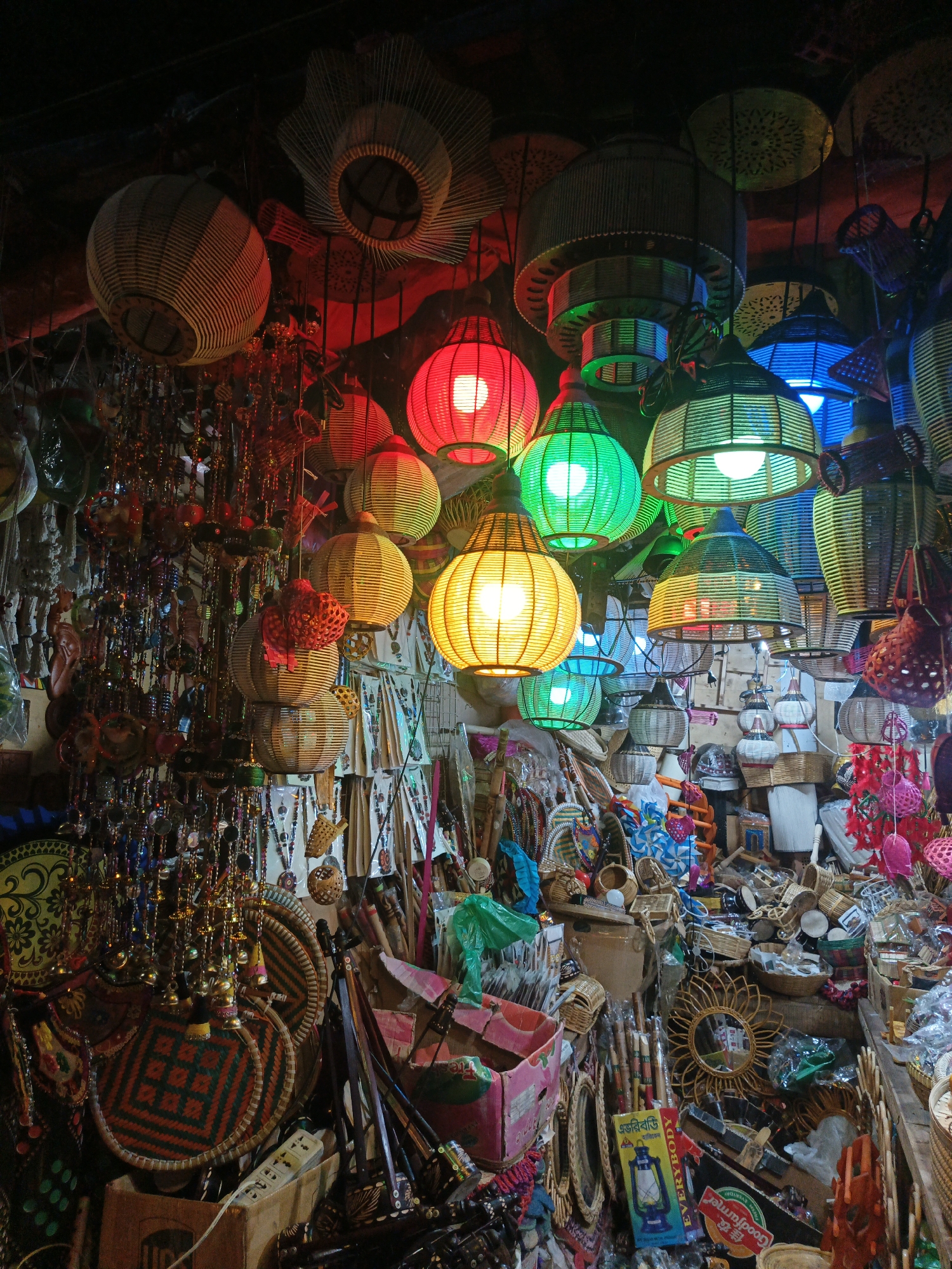 The image depicts a vibrant and densely packed artisan shop or market stall, likely indoors, in Dhaka, Bangladesh. The scene is illuminated by numerous hanging lanterns, primarily crafted from natural materials like wicker or bamboo, which cast a colorful glow throughout the space. These lanterns are diverse in shape—including round, conical, cylindrical, and star-like forms—and emit light in a spectrum of red, green, yellow, and blue hues.
The shop is overflowing with a wide array of handmade goods. Below the suspended lanterns, there are copious beaded curtains and hanging decorations, some featuring small bells or mirror accents. Woven baskets, intricately patterned trays, and fan-like items are stacked and displayed. Other visible objects include small mirrors with decorative frames, various wooden crafts, and an assortment of trinkets and textiles. The overall impression is one of rich texture and color, characteristic of a bustling craft market.
No people are visible, but the extensive display of goods points to a commercial setting. The artificial, multi-colored lighting, provided mainly by the lanterns, suggests it is either nighttime, a dark interior space, or a covered stall. The style of the crafts, especially the woven items and decorative lanterns, is consistent with regional craftsmanship found in South Asia.
Visible text includes "UPS" on a cardboard box. On a product package, Bengali script reads "একটি অপূর্ব সুন্দর লণ্ঠন" (A wonderfully beautiful lantern) and "এভারগ্রিন" (Evergreen). Another label partially visible says "GoodFarmer." The scene evokes a sense of cultural vibrancy and abundant craftsmanship.