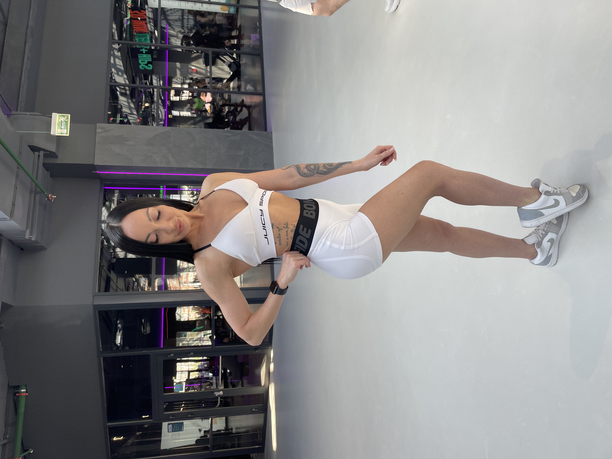 A woman is posing in a gym. She is wearing a white sports bra and shorts with black trim and grey and white athletic shoes. She has a tattoo on her left arm. The gym appears to be located in a modern building with large windows. The scene is inside a fitness center, possibly in Kazan, Russia.