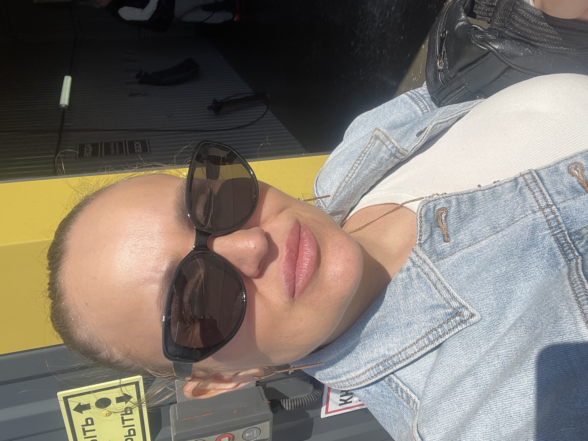 A woman wearing sunglasses and a denim jacket is positioned indoors, possibly in an industrial or commercial area in the city of Chaikovsky, Russia. A black bag is visible next to her. A sign in Russian is partially visible in the foreground, attached to a piece of equipment.