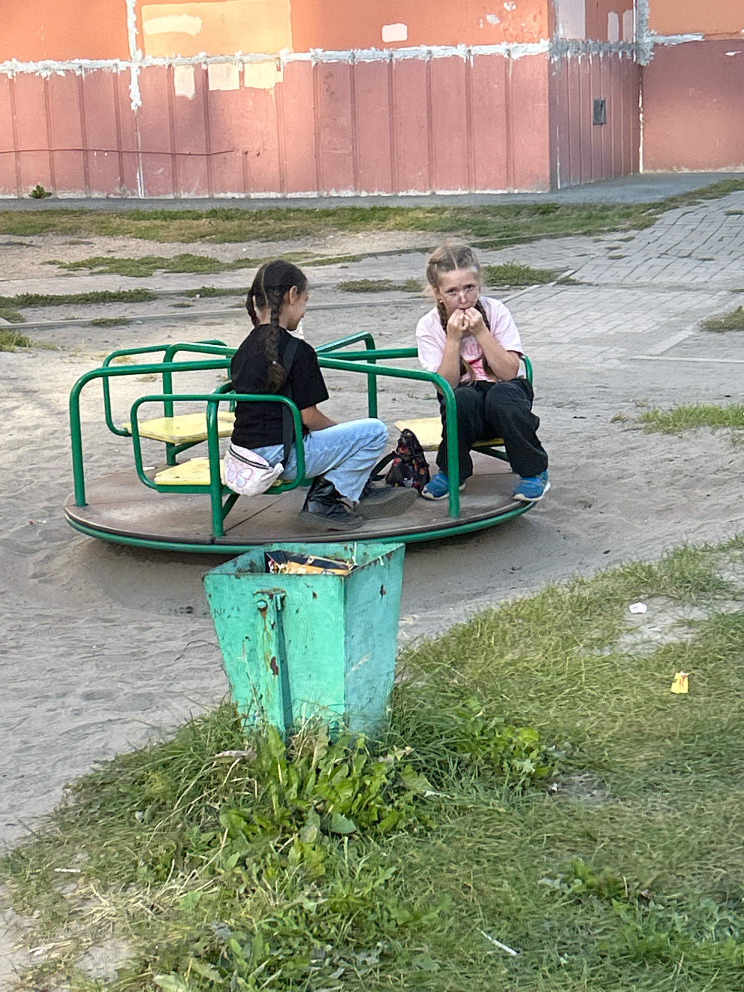 Two young girls are on a merry-go-round in a playground. The girl on the left, with dark hair in a braid, wears a black t-shirt and blue jeans, and has a light-colored fanny pack around her waist. The girl on the right, with braided blonde hair and glasses, wears a pink t-shirt and dark pants, and is biting her fingernails. The merry-go-round is green with yellow seats. Next to the merry-go-round is a green metal trash can filled with trash. The playground is covered in sand and patches of grass. In the background, there is an orange building with a tiled facade. The scene appears to be in Tomsk, Russia, based on the architectural style of the building and the type of playground equipment.