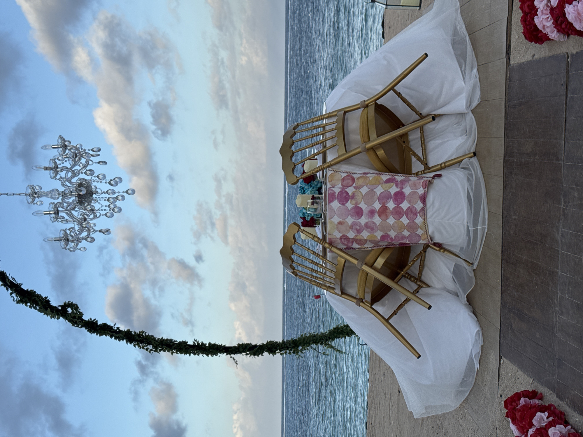 A horizontally oriented, medium shot captures an outdoor wedding setup. The background reveals the ocean and sky, exhibiting white clouds against a blue backdrop.
In the foreground, two gold chairs are arranged at a table draped in white fabric. A patterned cushion adorns one of the chairs. Suspended above the table is an ornate chandelier, partially obscured by foliage.
Red and white floral arrangements are positioned at the bottom right and upper right corners of the frame.
Based on the location of the image and details such as the ocean, sky, wedding setup, and style of the furniture, it is likely this photo was taken in the coastal district of Kuta Selatan, Indonesia. However, the specific venue cannot be determined from the image alone.