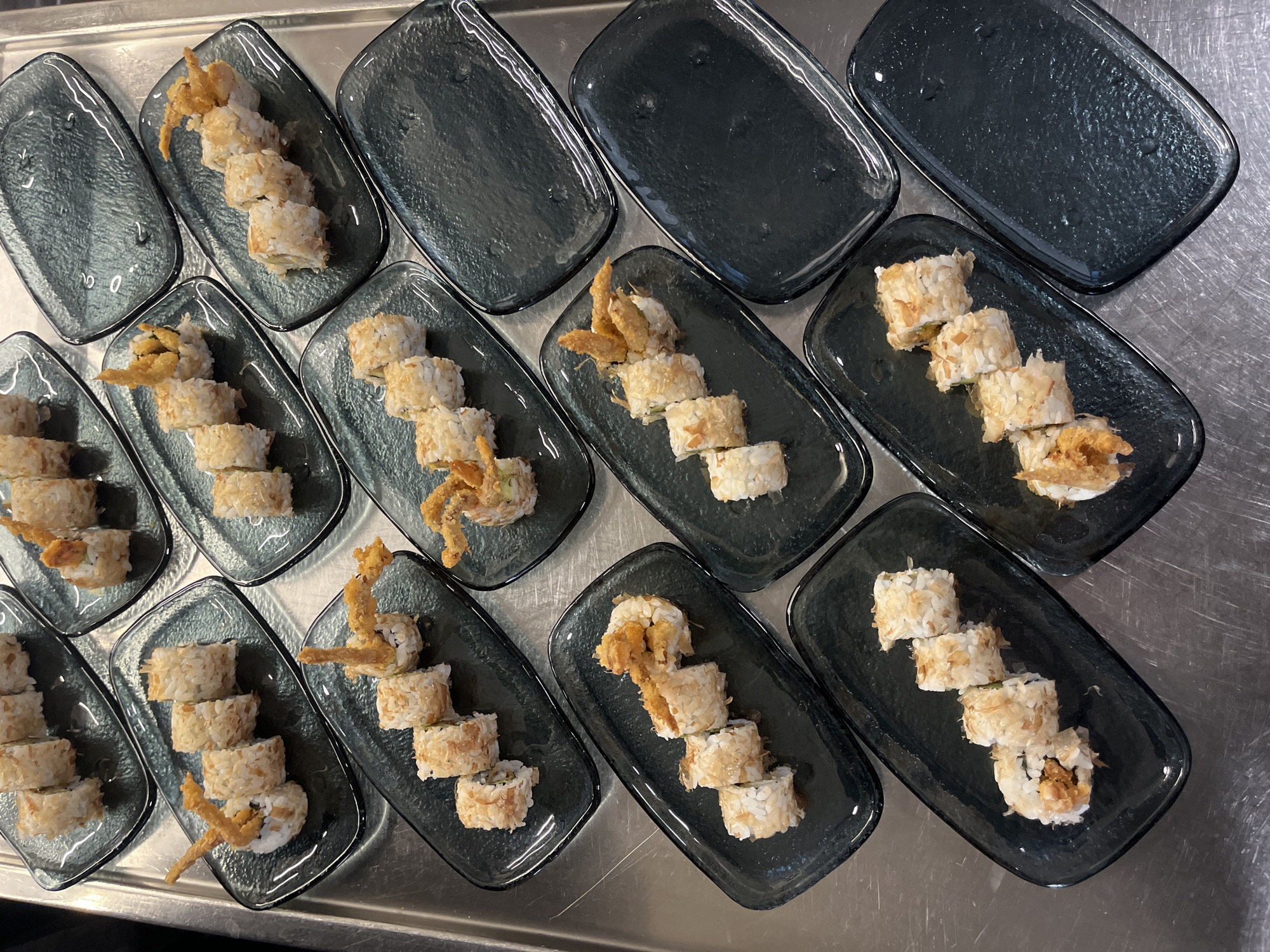 The image shows a top-down view of twelve small, dark gray glass plates arranged on a stainless steel surface. Each plate contains sushi rolls. Some plates have sushi rolls topped with fried crab. The arrangement suggests a catered event or a banquet setting. There are no people visible in the image. The location in Sofia, Bulgaria cannot be determined from the image.