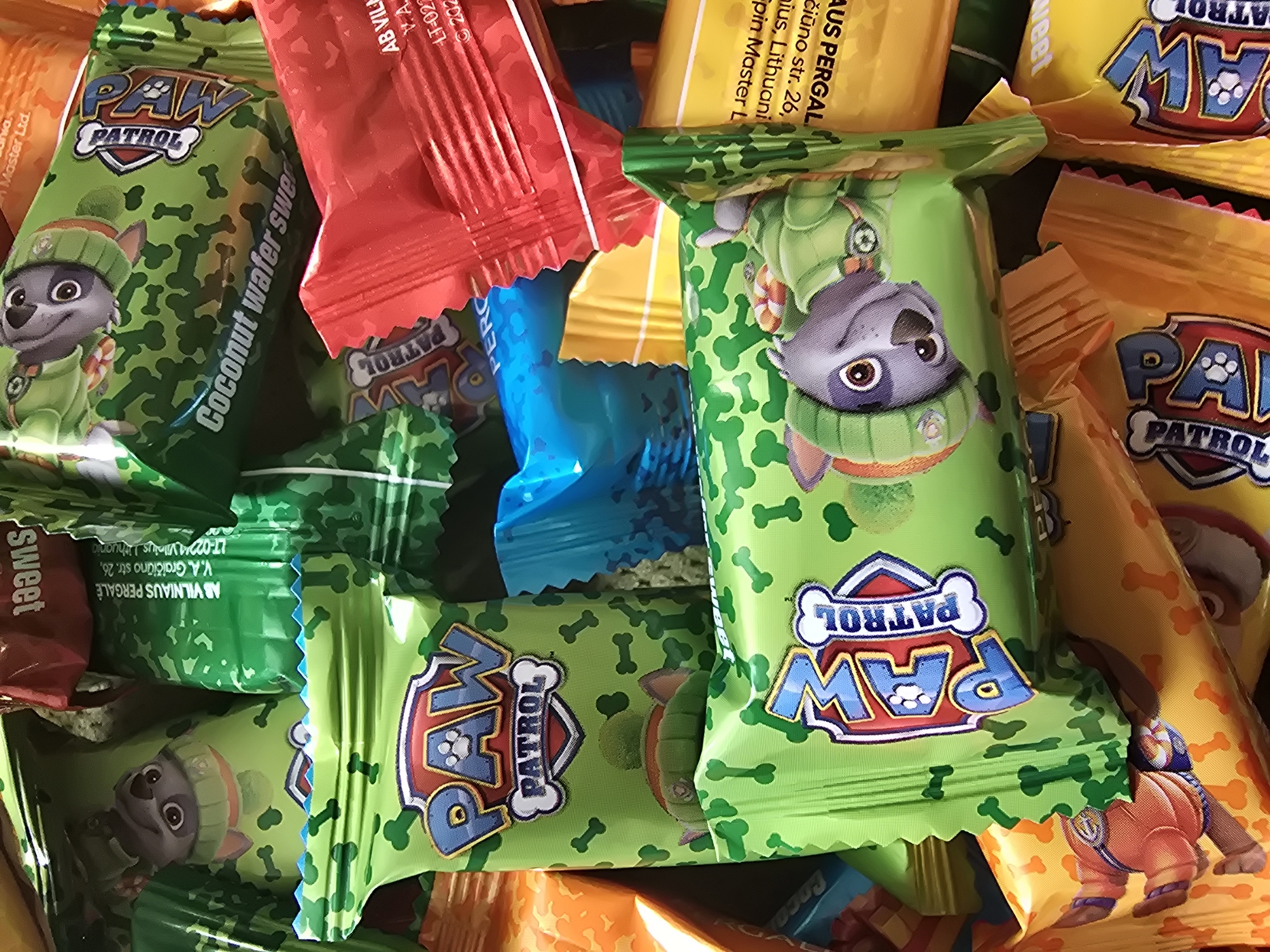 The image shows a close-up, high-angle view of a pile of individually wrapped Paw Patrol-themed coconut wafer sweets. The wrappers are colorful, primarily green, orange, yellow, red, and blue, each featuring Paw Patrol characters and the "Paw Patrol" logo. The sweets appear to be piled haphazardly, with some overlapping others. Text printed on some of the wrappers includes "Coconut wafer sweet," and the address "V.A. Graičiūno str. 26, LT-0224 Vilnius, Lithuania."