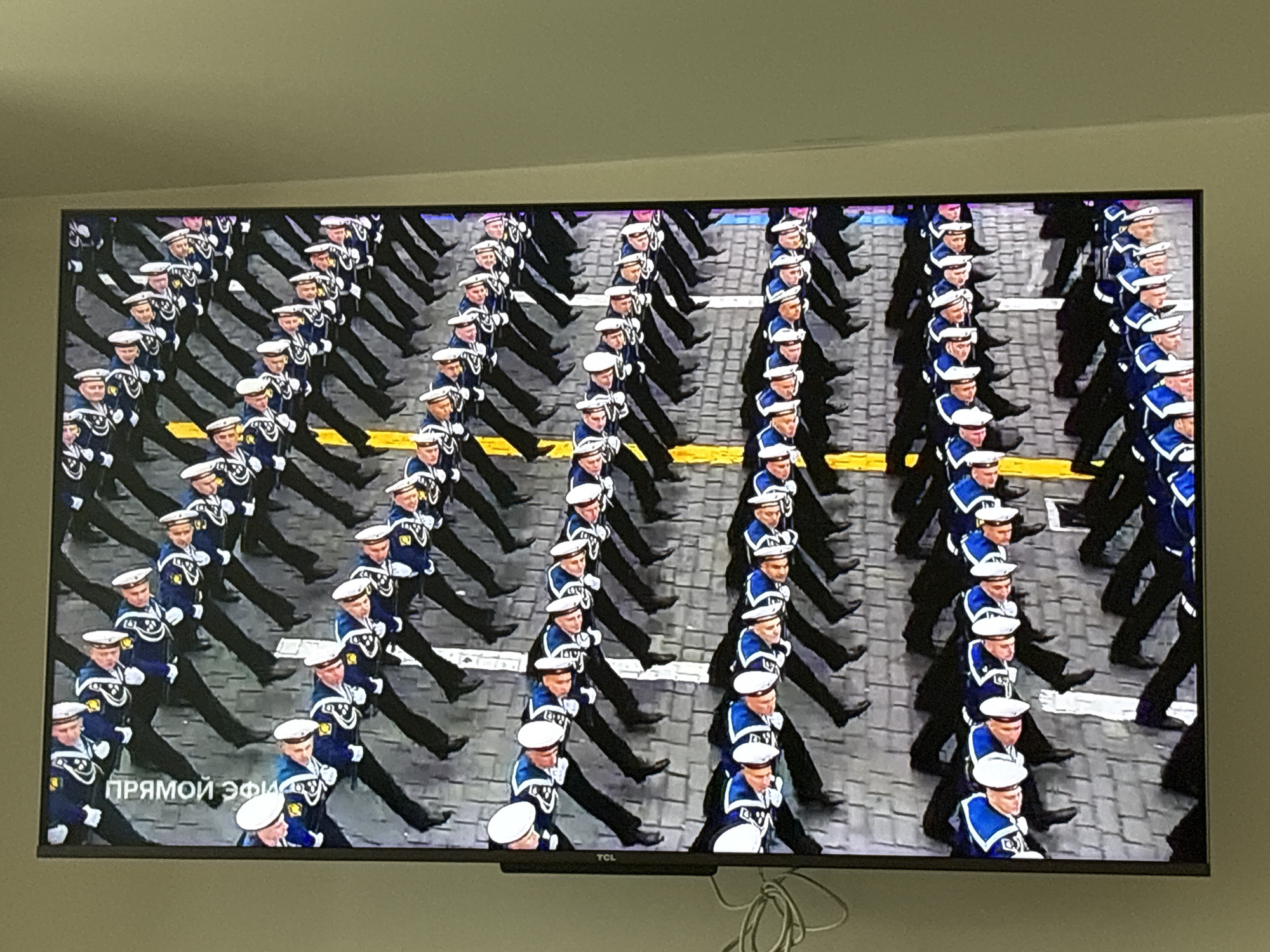 A large group of Russian Navy sailors are marching in formation on a paved surface. They are wearing navy blue uniforms with white caps, and are arranged in neat rows and columns. The location is likely a city square in Belokurikha, Russia, although this cannot be definitively determined from the image. The text "Прямой эфир" is visible in the lower left corner of the screen, indicating a live broadcast. The image is being displayed on a TCL television.