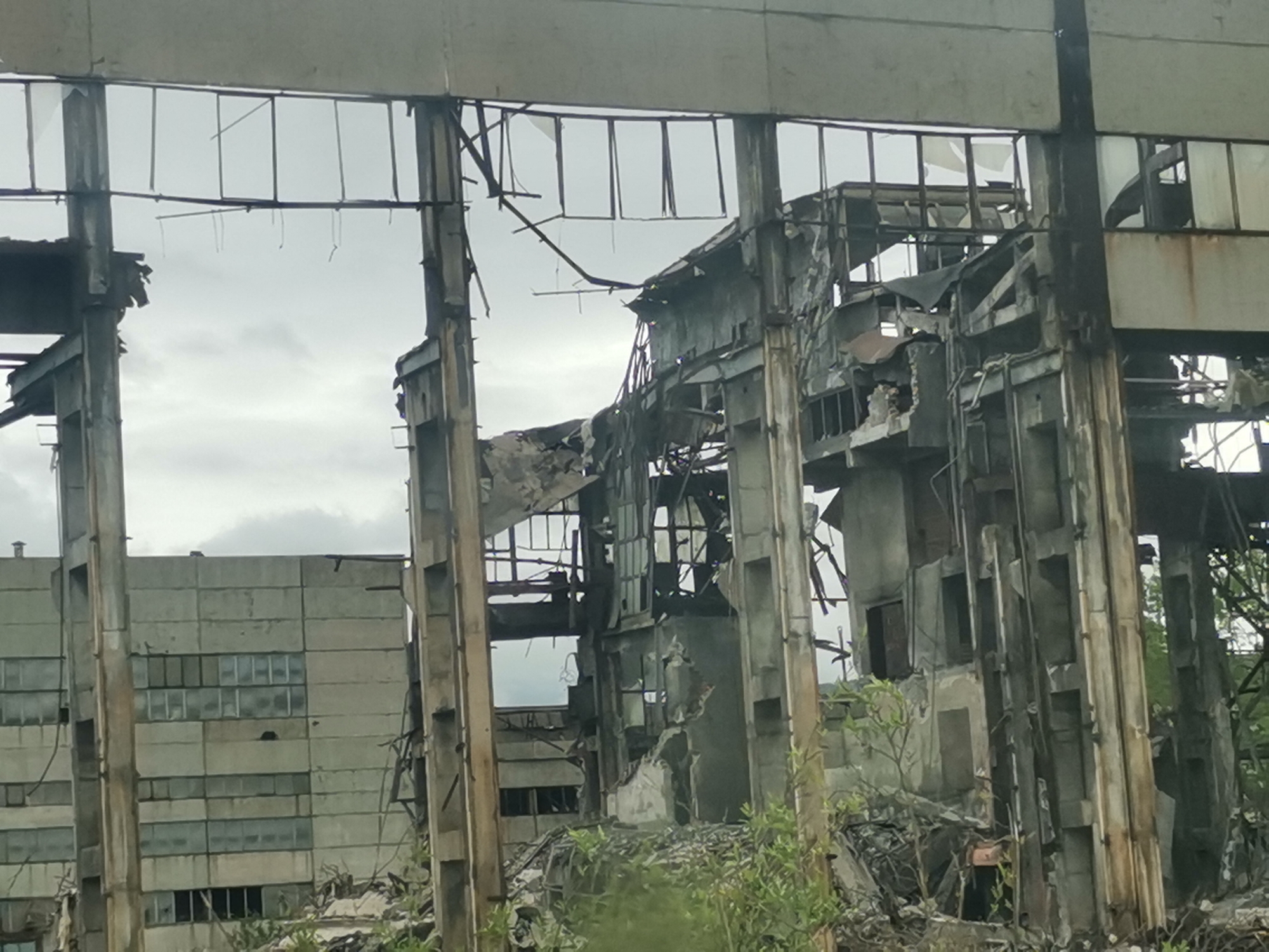 The image shows the ruins of a large industrial building in what appears to be a state of significant disrepair, possibly located in Zhivkovo, Bulgaria. The building is made of concrete and steel, with large sections collapsed or destroyed. Parts of the frame and some walls remain standing. Rubble and debris are visible on the ground. Another concrete building with intact windows is visible in the background. Vegetation is growing in the foreground, suggesting that the building has been abandoned for some time. The sky is overcast. There are no people visible in the image.