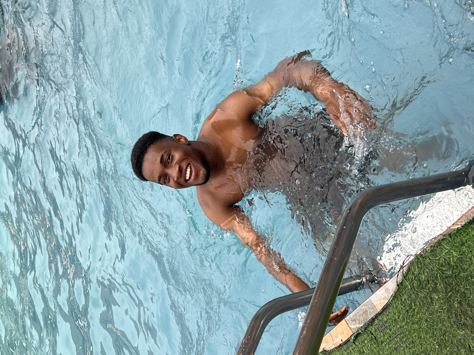 A young, dark-skinned man is captured smiling broadly and looking directly at the camera while swimming in a clear blue pool. His upper torso and head are visible above the water, which shows ripples and splashes from movement. He is shirtless, with wet skin and short, dark hair. His right arm appears to be near or holding onto a metallic pool ladder, which is visible in the foreground along the edge of the pool. Next to the pool's edge, a strip of artificial green grass is present, suggesting a recreational setting. The lighting indicates it is daytime. The man's joyful expression conveys a sense of relaxation and enjoyment. The scene depicts a casual moment of swimming, likely in Nkpor, Nigeria. No visible text is present.