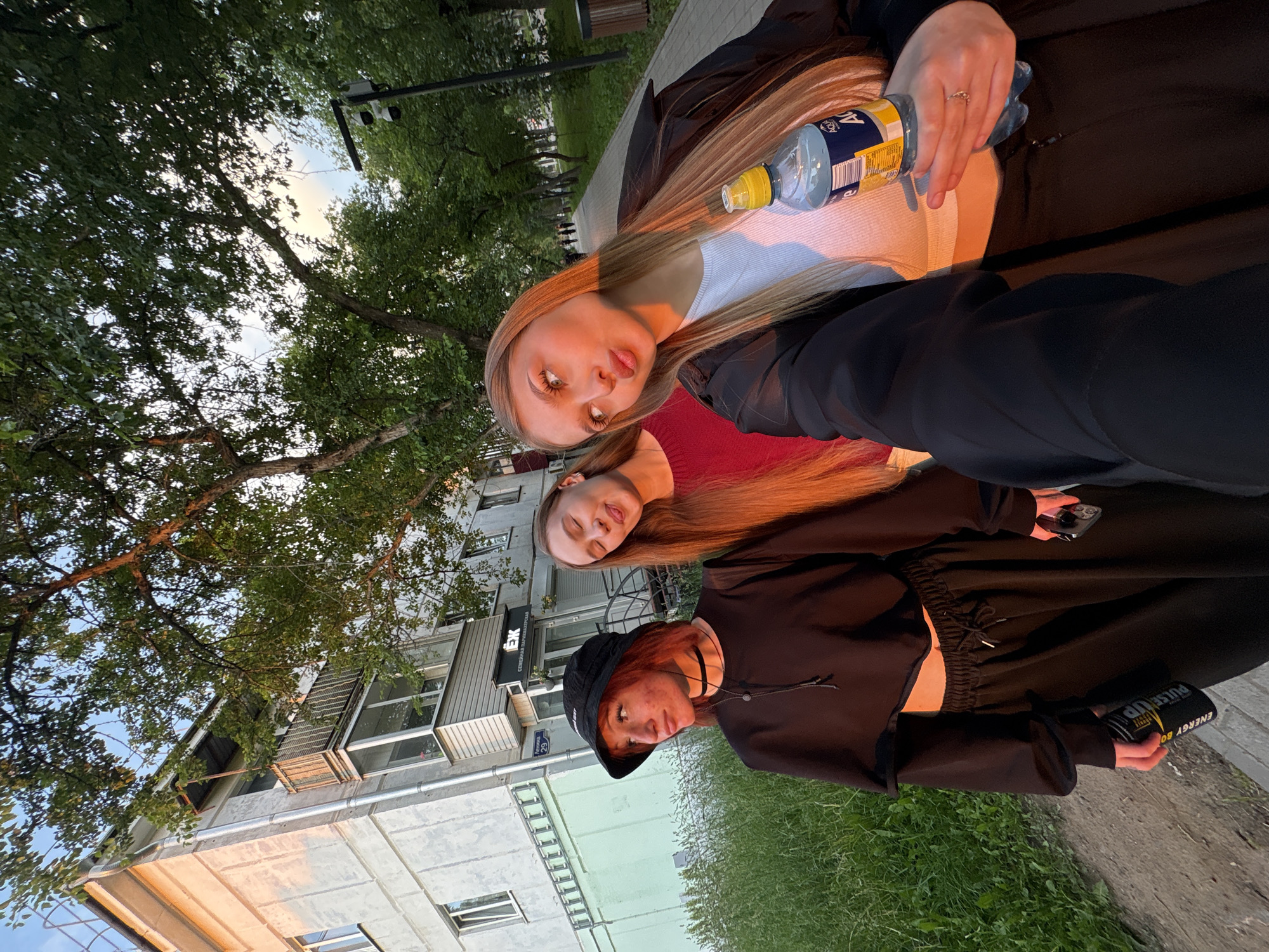 Three young women pose for a selfie outdoors in Chaikovsky, Russia. The woman on the left wears a black bucket hat, a black crop top, and black pants. She has red hair and is wearing a black choker. The woman in the middle has long red hair and wears a red shirt. The woman on the right has long blonde hair and wears a white top under a black jacket. She holds a bottle of water. The background shows a building with a balcony, trees, and a paved walkway. The sun appears to be setting, casting a warm glow on the scene. The building has an "EX" sign in Russian above the entrance.