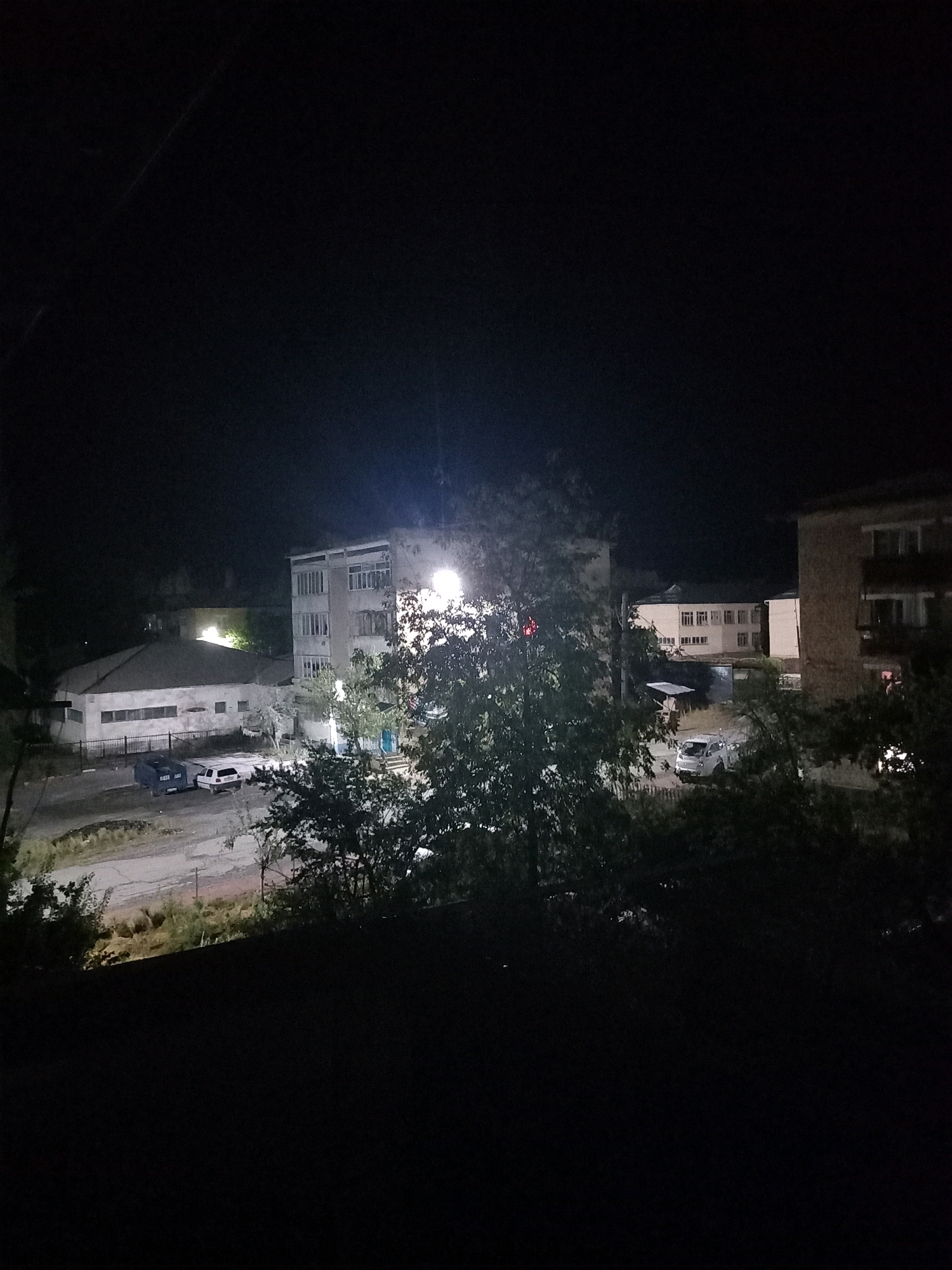 A nighttime, outdoor, elevated shot captures a street scene in Aydarken, Kyrgyzstan. The image features several buildings, including a multi-story concrete structure on the right side, illuminated by a bright overhead light, and a long, single-story building to its left. A white car is parked near the single-story building, and two large dark bins are situated next to it. In the distance, another building with multiple windows is visible. Trees with dense foliage are scattered throughout the scene, with some partially obscuring the view of the buildings. The overall lighting is dim, with artificial lights from the buildings and street providing illumination. The ground appears to be paved with asphalt or concrete in some areas, and dirt or gravel in others. The sky is completely dark, indicating it is nighttime.