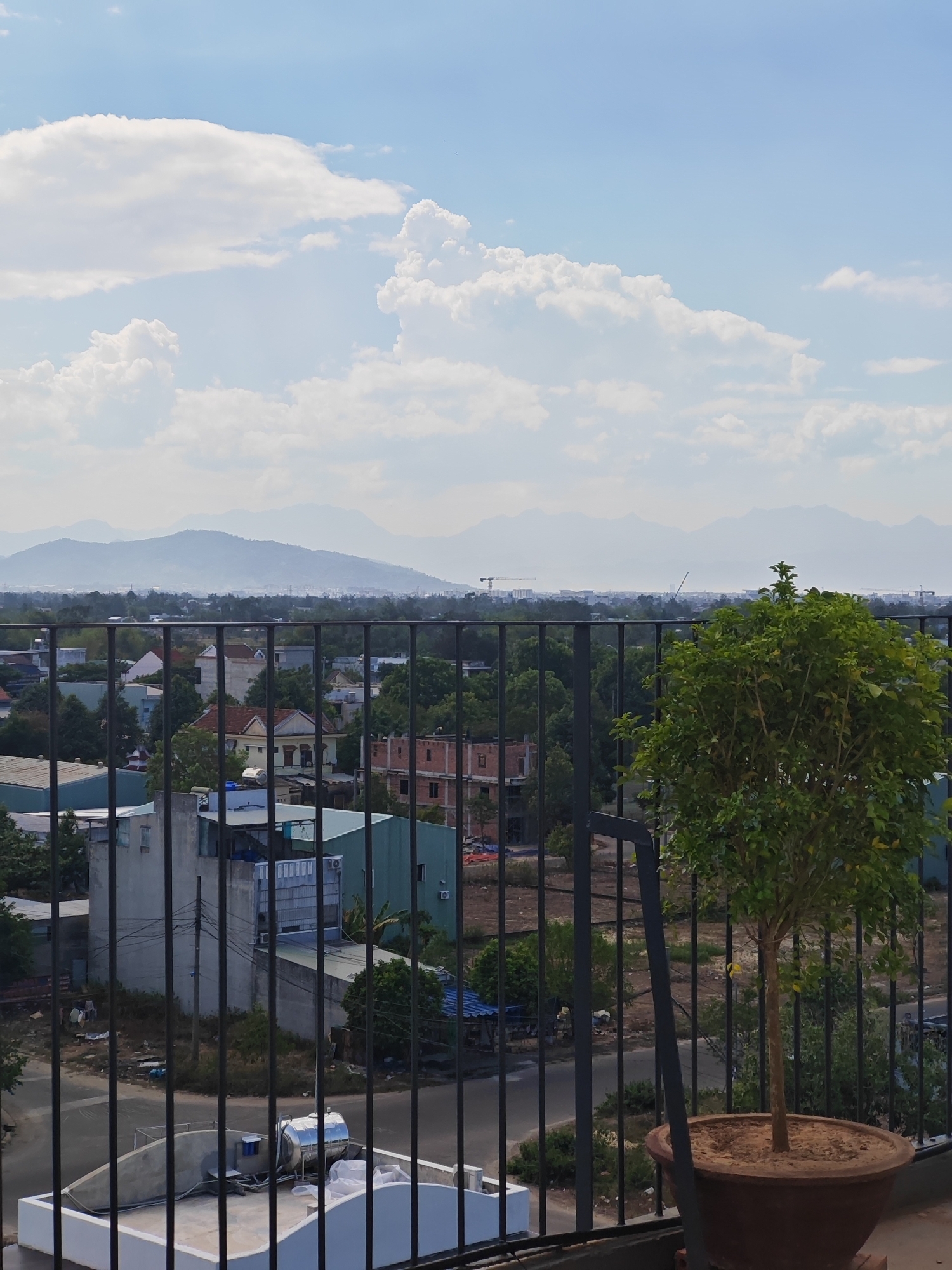 The image captures an aerial view of a suburban area in Đà Nẵng, Vietnam, on a bright, partly cloudy day. In the foreground, a black metal fence obstructs the view of a rooftop with a large silver water tank and some white tarps. To the right of the fence, a potted tree with green foliage stands prominently. Beyond the fence, a dense canopy of green trees forms the midground, punctuated by various residential buildings with tiled roofs and some under construction. In the distance, a hazy range of mountains stretches across the horizon under a pale blue sky filled with scattered white clouds. The scene suggests a developing urban environment where residential development is interspersed with natural greenery and the natural landscape.