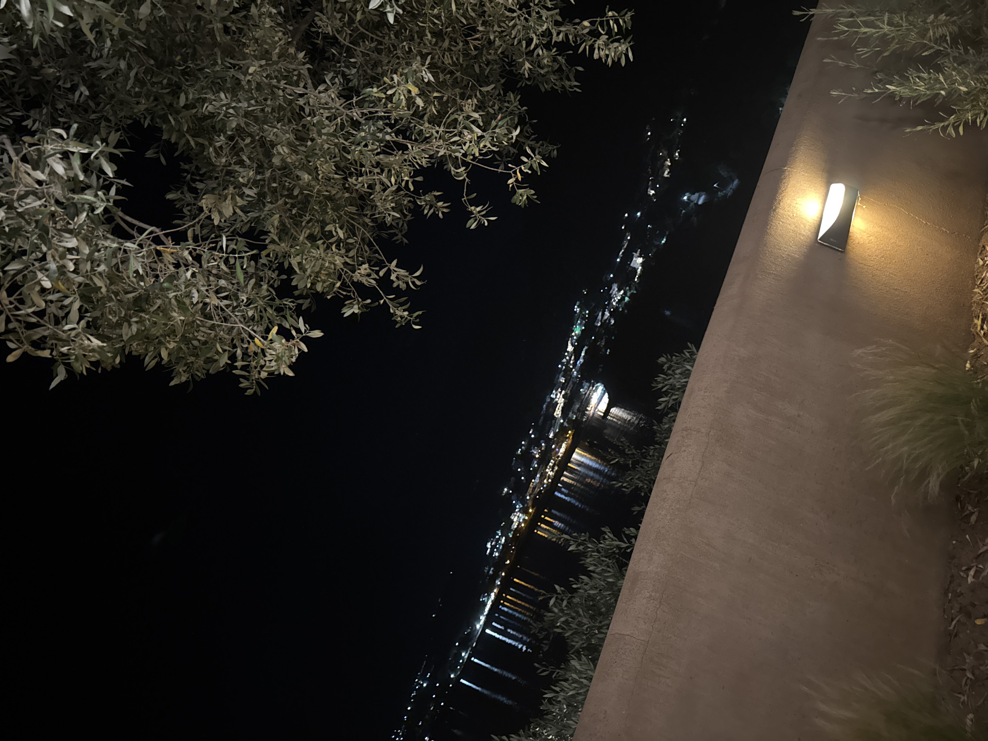 The image presents a nighttime scene of Golden Beach, Greece, captured from an elevated perspective. In the foreground, a prominent reddish-brown textured wall extends horizontally across the lower third of the frame. A modern, rectangular wall-mounted light fixture, emitting a warm yellow glow both upward and downward, is positioned on this wall in the lower right quadrant. At the base of the wall, sparse vegetation, including light-colored grasses and small leafy shrubs, is visible.
Above the wall, a dark body of water, likely a bay, occupies the midground, reflecting numerous artificial lights from a distant shoreline. These reflections appear as distinct, elongated vertical streaks of light on the water's surface. In the background, a coastal town or settlement is discernible by its scattered artificial lights, outlining buildings and infrastructure against the dark landscape. The upper right portion of the image is filled with the dense, light-colored foliage and branches of a large tree, consistent with an olive tree, partially obscuring the dark, unlit night sky. The overall scene is dark, punctuated by the various artificial light sources. No individuals, direct actions, or specific interactions are depicted. The image itself exhibits a slight upward diagonal tilt from left to right.