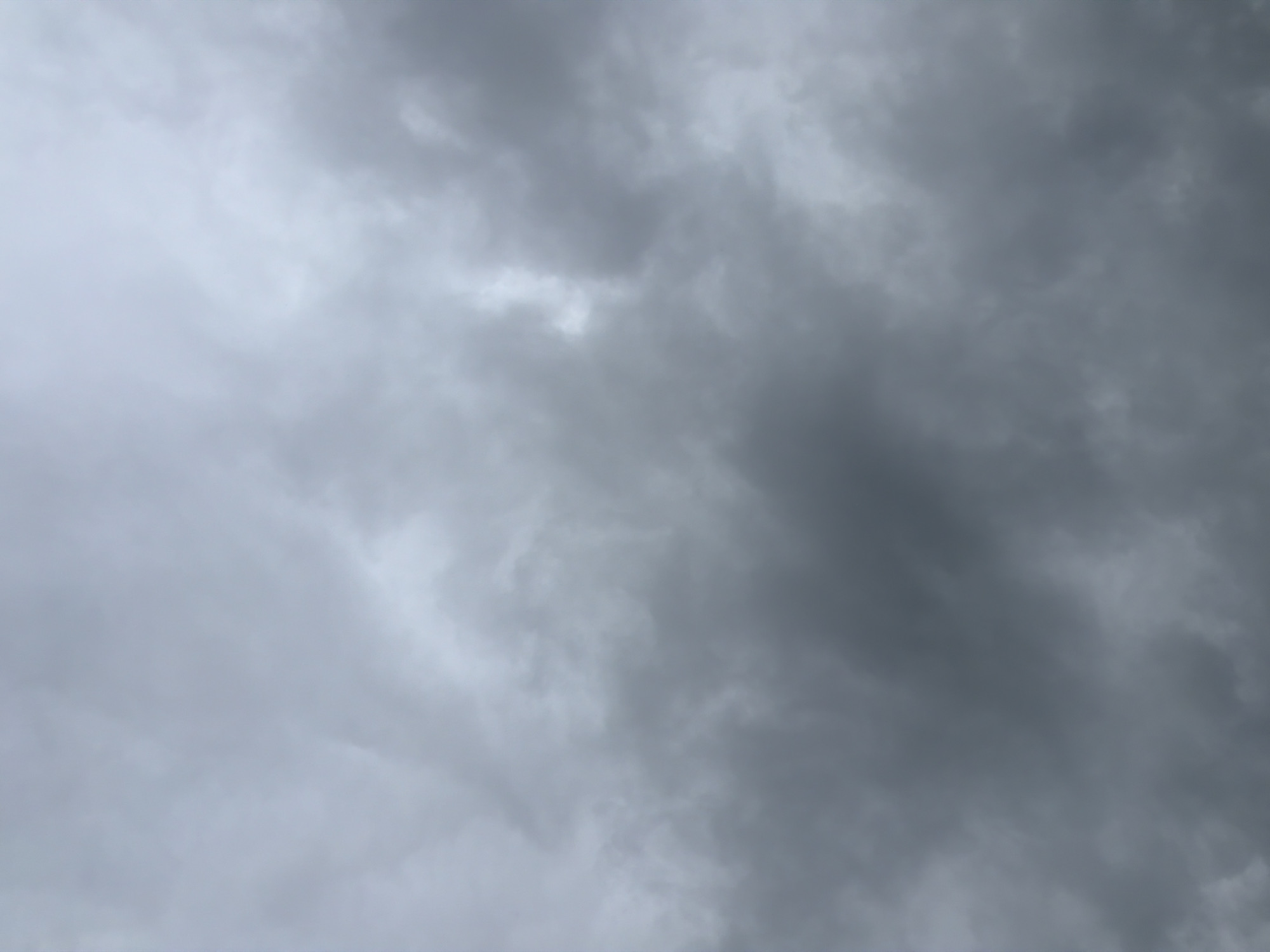 The image is a close-up, overcast sky. The clouds are a uniform grey, with some areas appearing darker and denser than others, suggesting a heavy cloud cover. There is no visible sun or clear sky. The scene is devoid of any objects, people, or discernible features, focusing solely on the texture and tonal variations of the clouds. The location in Męcina, Poland, is not visually represented in the image itself; it is contextual information provided separately.