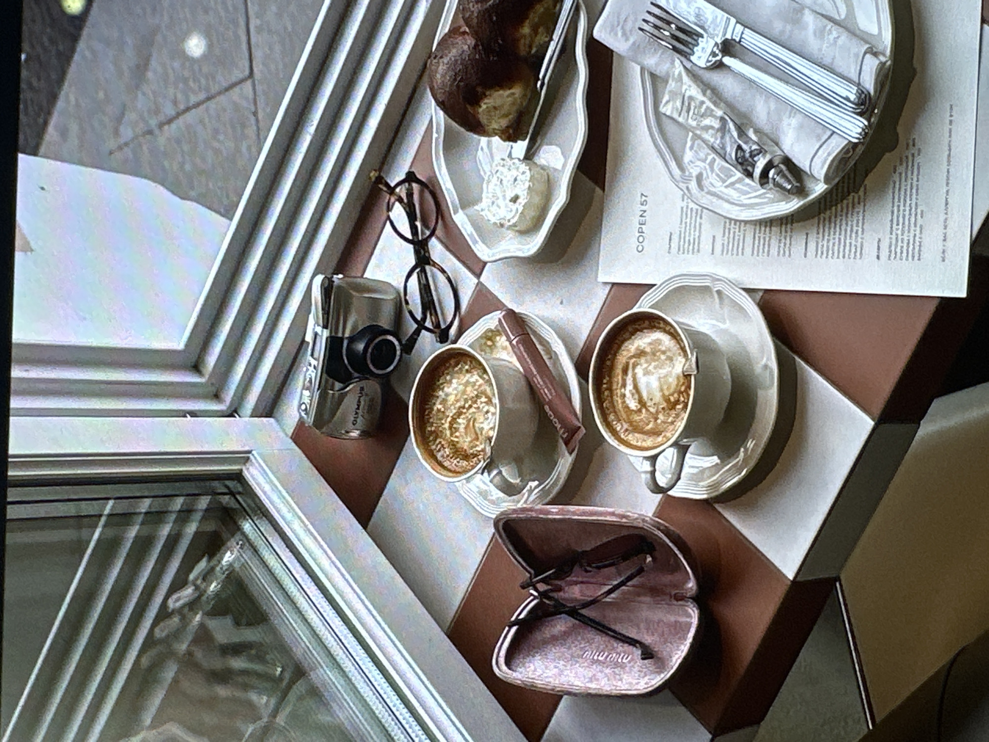 A high-angle shot shows a tabletop arrangement on a checkered brown and white surface, positioned near a window with a visible view of the exterior in Yerevan, Armenia. Two cups of coffee with latte art sit on saucers, accompanied by a small brown tube. An open eyeglasses case revealing a pair of eyeglasses labeled "miu miu" rests on the table. Also present are a pair of eyeglasses outside their case, a compact Olympus camera, a plate of pastry with butter, and a menu item labeled "COPEN 57." Cutlery wrapped in a napkin is positioned atop the menu. The scene appears to be set in a cafe or restaurant.