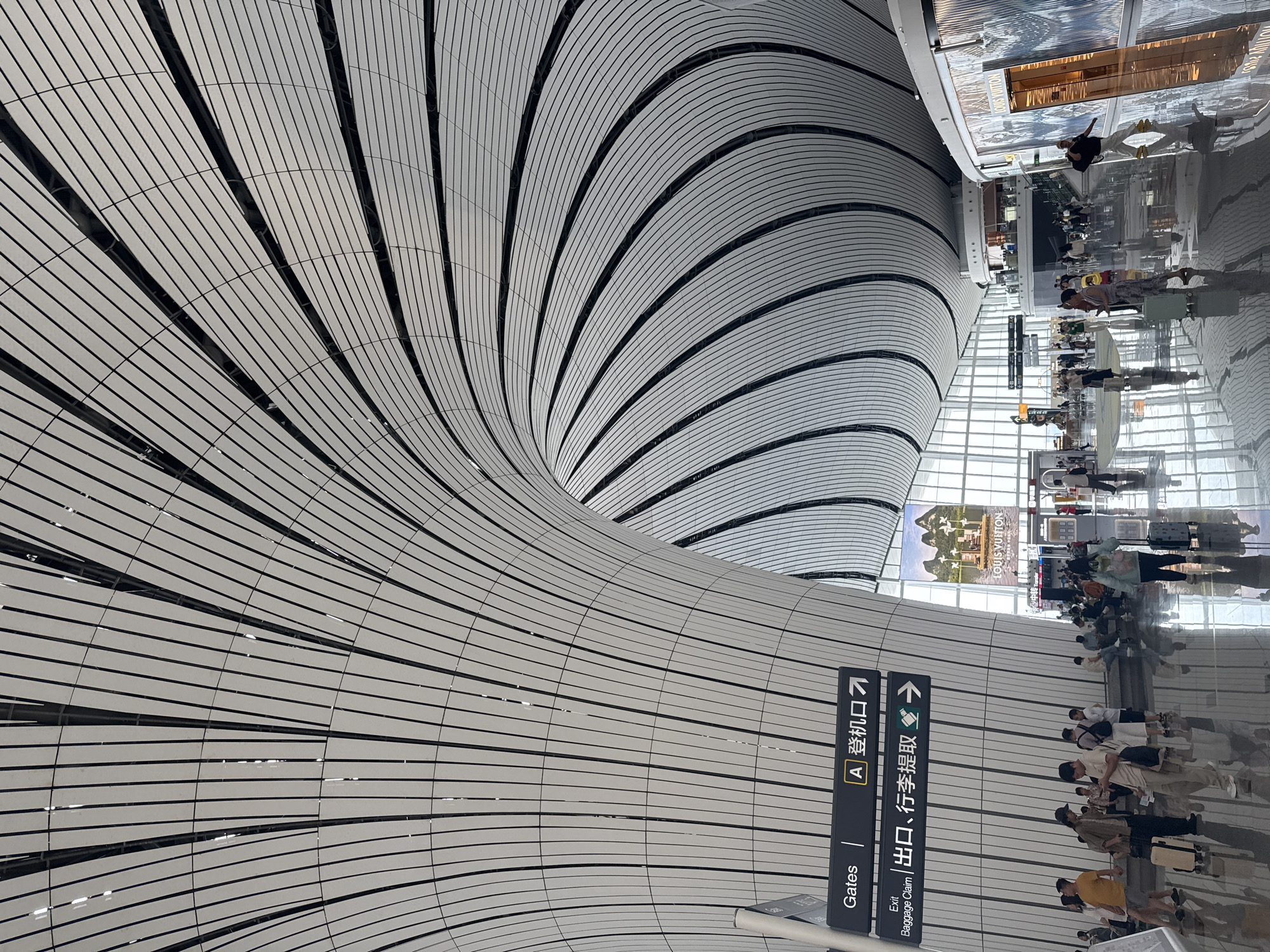 This image depicts the interior of Beijing Daxing International Airport in Guangyang District, China. The airport's distinctive architecture features a futuristic, flowing roof structure composed of white panels with black trim, creating a dramatic, curved canopy overhead. In the foreground, people are seen walking and standing, some with luggage, reflecting on the polished, glossy floor. Signage indicates "Gates" and directions to "A 登机口" (A Boarding Gate) and "Exit Baggage Claim 出口、行李提取" (Exit, Baggage Claim). A large advertisement for Louis Vuitton is visible in the background, suggesting a retail area within the airport. The overall atmosphere is one of a modern, bustling transportation hub.