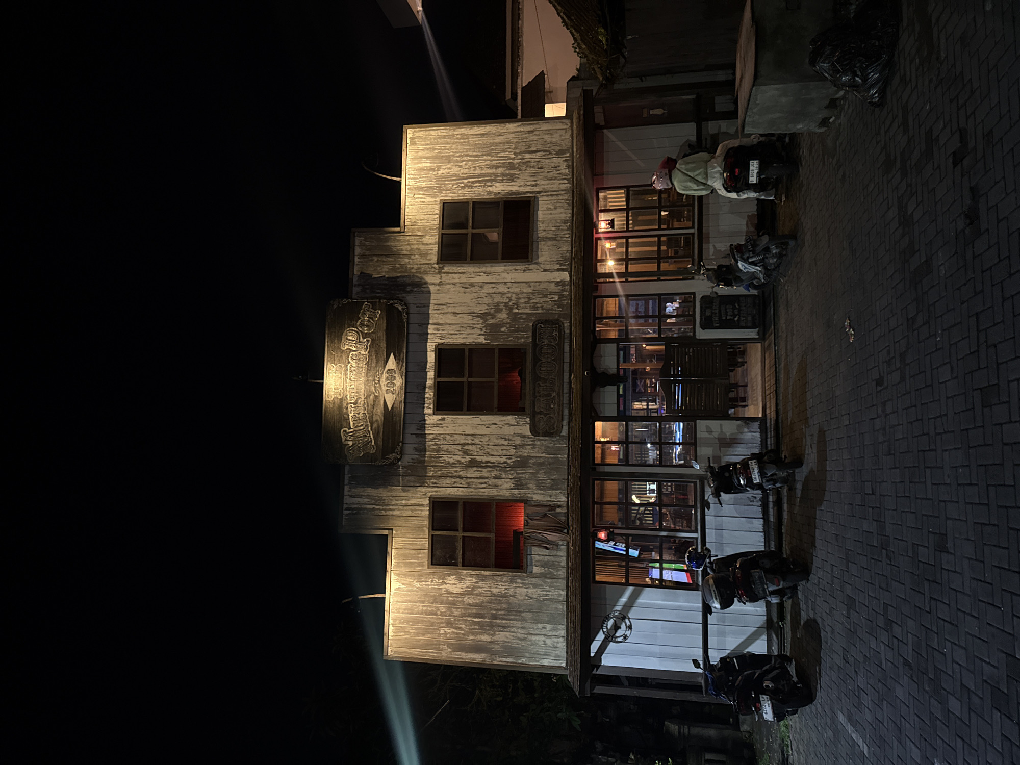 At night in Ubud District, Indonesia, a three-story building with a weathered wood facade stands brightly lit. The building resembles an old western-style structure, possibly a bar or restaurant, evident from the signage above the door. The facade features multiple windows with warm light emanating from within. Several scooters are parked along the sidewalk in front of the building. A person wearing a light-colored jacket and helmet is near the scooters closest to the building. Trash bags are visible at the edge of the sidewalk and street. The sky is dark and clear above the building.