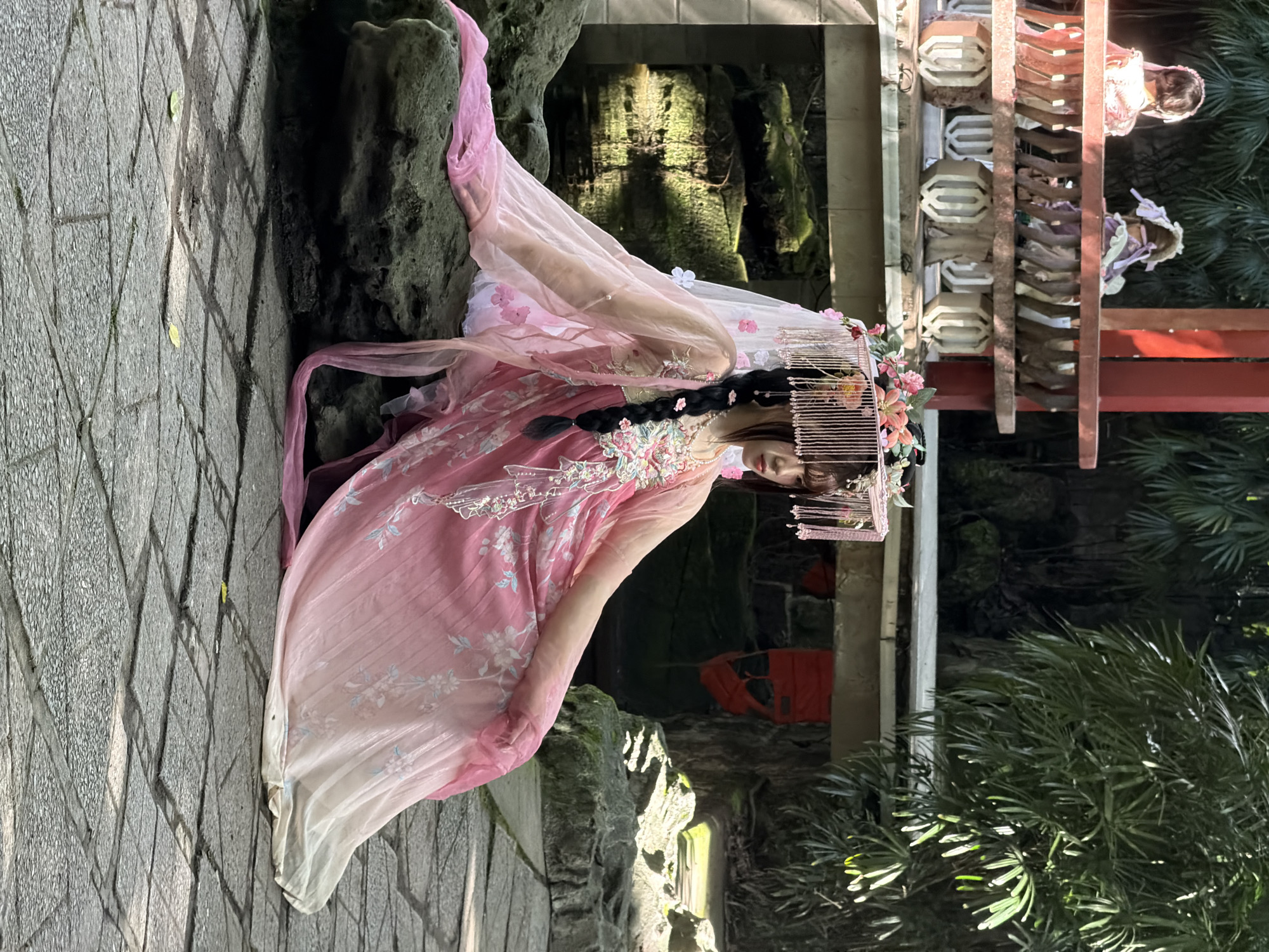 A young woman with dark hair in a braid, wearing an ornate pink and white traditional Chinese dress and a decorative headpiece adorned with flowers and beadwork, is seated on a large, moss-covered rock in a garden. The dress features flowing sleeves and a gradient of pink to white, with floral embroidery. She is looking to her right with a serene expression. Behind her, a wooden balcony structure with carved details is visible, and in the background, lush green foliage and a rocky outcrop with moss are present. The ground is paved with square stone tiles. The overall scene evokes a sense of traditional aesthetics and tranquility. The location is identified as Yuzhong District, China.
