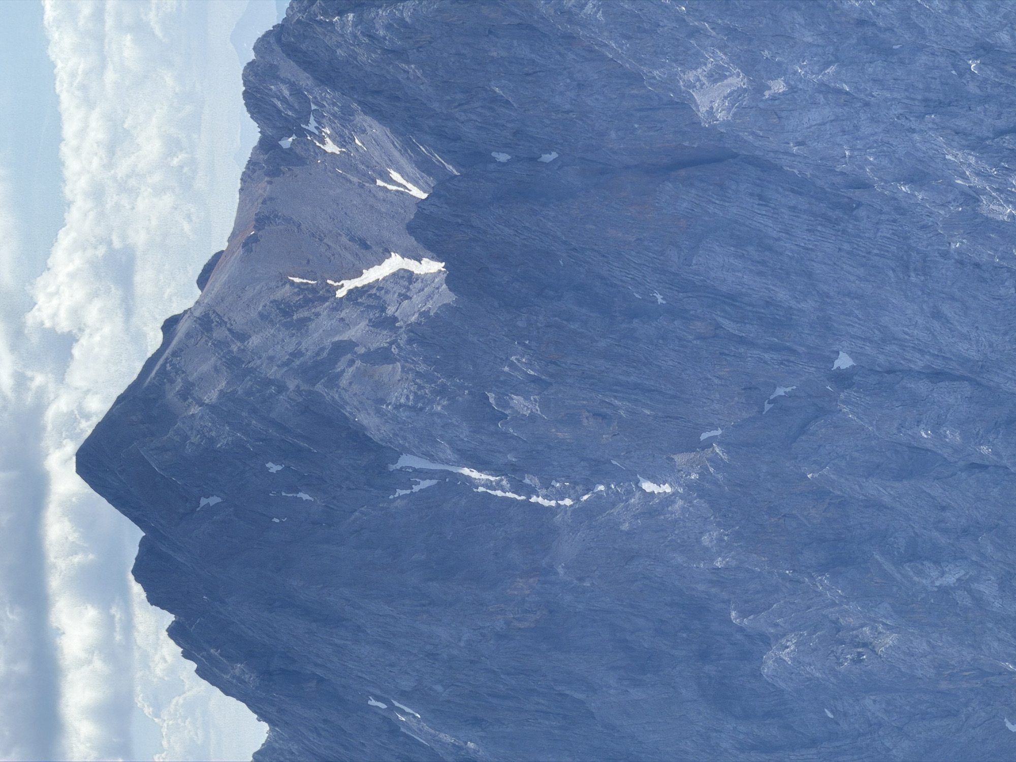 This is a close-up, high-angle aerial photograph of the Zugspitze, the highest peak in Germany, located in the Wetterstein Mountains near Garmisch-Partenkirchen. The mountain is composed of massive, rugged rock faces with steep inclines. Patches of snow and ice are visible in ravines and on upper slopes, particularly in a prominent channel running down the central face. The rock surface is textured with numerous striations and crevices, indicating significant erosion. The sky above is partly cloudy, with distinct bands of white clouds against a pale blue backdrop. The lighting suggests daytime, with the sun casting subtle shadows that highlight the mountain's topographical features. No people or man-made structures are visible in this extreme close-up of the natural landscape.