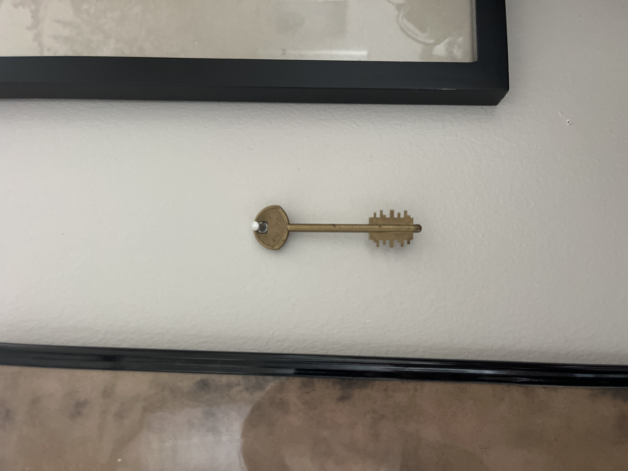 The image shows a vintage-style brass key hanging from a nail on a white wall. The key has a rounded head and a toothed bit. The wall appears to be in a home or office setting. Above the key, a portion of a black picture frame with a light-colored image inside is visible. Below the key, another black picture frame is partially visible. The lighting suggests an indoor environment, likely during the daytime. No people are visible in the image. The overall impression is one of simple, decorative display.