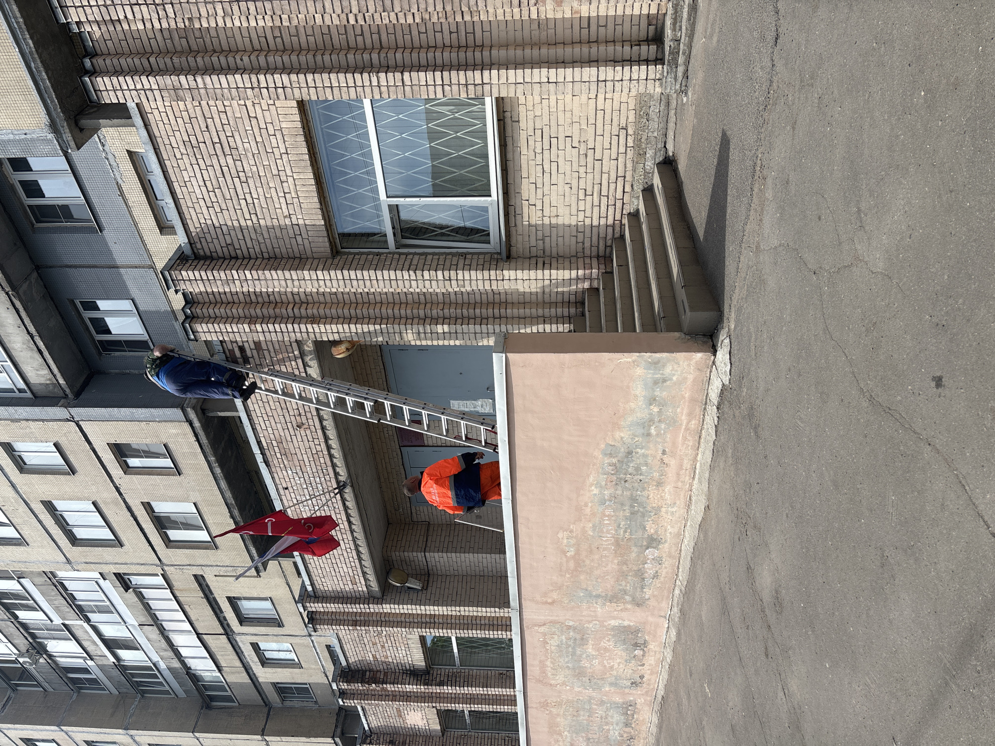 The image shows two people on a ladder repairing a building. The building is made of brick and has a window with a metal grate. The ladder is leaning against the building, and the two people are climbing up and down it. One person is wearing a blue jacket, and the other is wearing an orange jacket. There are two flags hanging from the building. The building is located in Saint Petersburg, Russia.