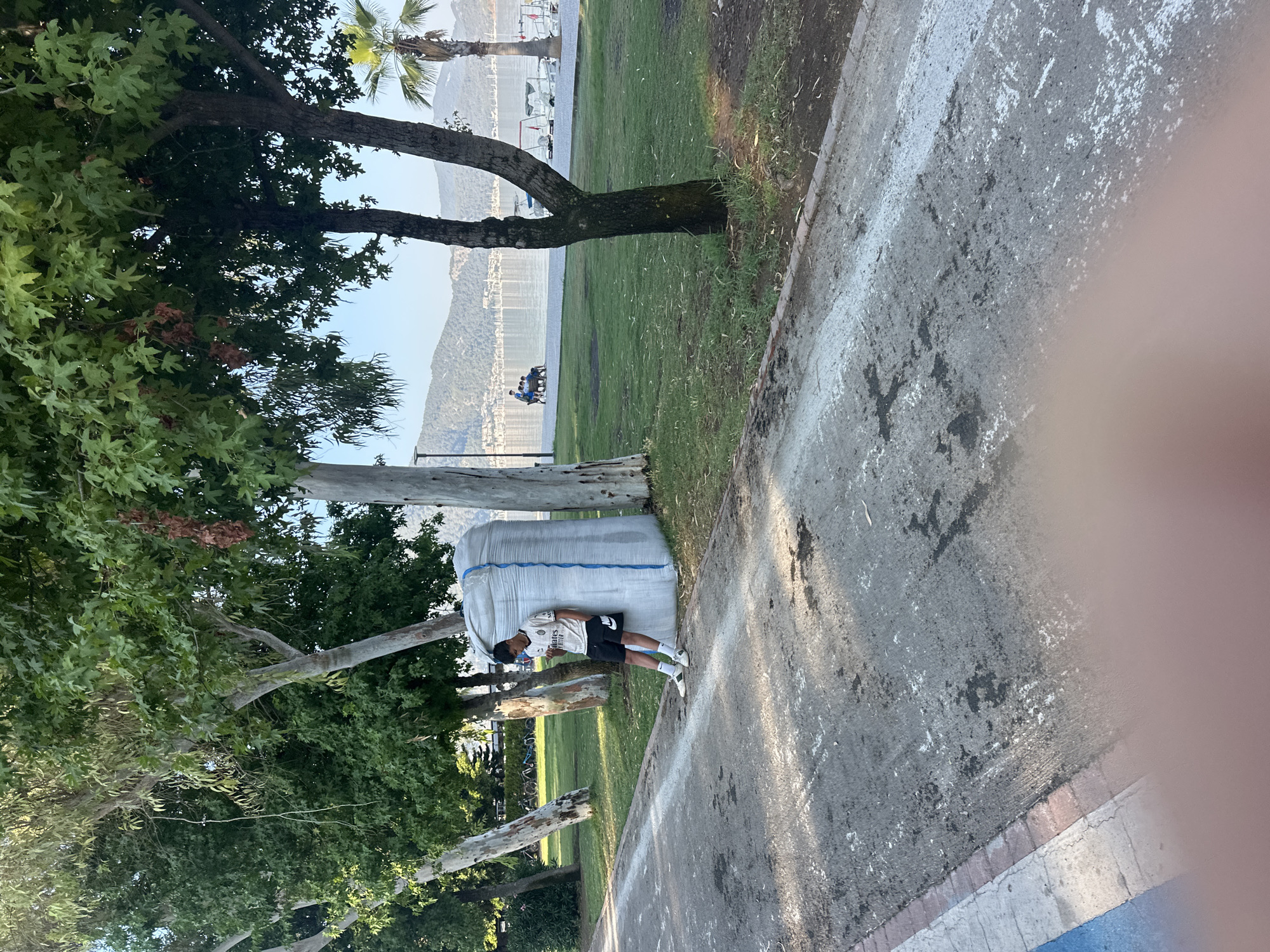 A young male is standing next to a large white sack, leaning against it and looking down at his phone. He is wearing a white soccer jersey with black trim and shorts, white socks pulled up to his knees, and white sneakers. The sack is tied with a blue rope. The scene takes place outdoors in a park-like setting. A paved pathway runs in the foreground, with a grassy area to the right. In the background, there is a body of water with several boats docked, and hills or mountains can be seen in the distance. The location is Fethiye, Turkey, likely along the Kordon, a popular waterfront promenade. The overall atmosphere is casual and relaxed.