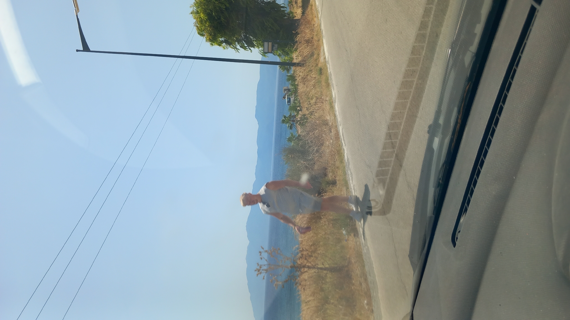 A man with blonde hair is walking on the side of a road next to the sea in Kos, Greece. He is wearing a light-colored sleeveless shirt and shorts. He is holding a red object in his left hand. The sea is visible behind him, with mountains in the distance. There is a street lamp on the right side of the image and power lines in the sky. The image appears to be taken from inside a vehicle, as indicated by the reflection on the windshield and the dashboard in the foreground.
