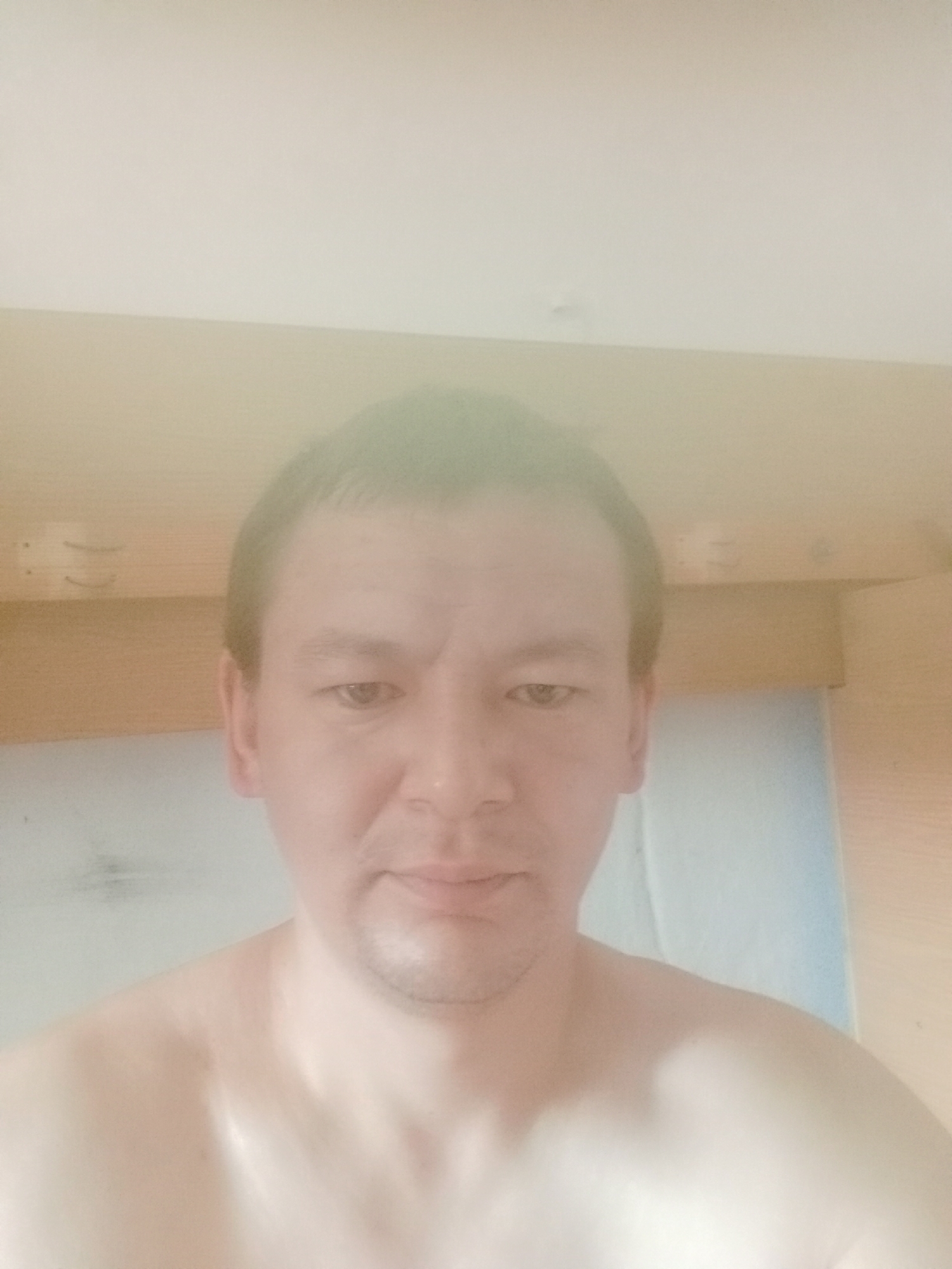 A close-up, head-and-shoulders selfie shows a fair-skinned man with light brown hair. He is looking directly at the camera with a neutral expression. His shoulders and upper chest are visible, and he appears to be shirtless. The background consists of a light-colored wall above and a wooden surface below. The image is overexposed and slightly blurry, making it difficult to discern fine details. There is no discernible indication of the location within the city of Aksu, Kazakhstan.