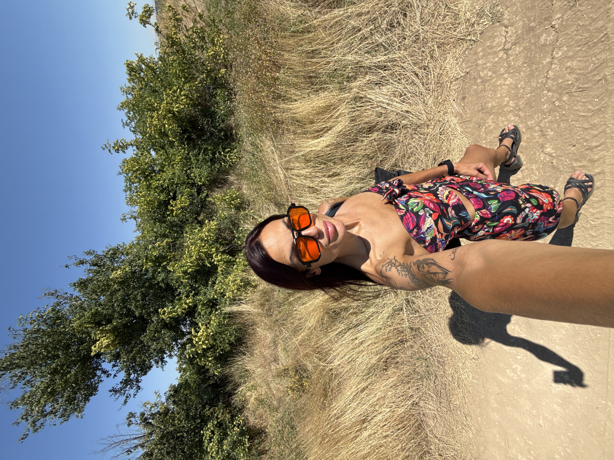 The image is a first-person perspective selfie taken outdoors on a sunny day. The foreground shows a person's arm holding a phone, casting a shadow on a dirt path. The person in the selfie is a woman with dark hair and sunglasses, wearing a colorful strapless top and a matching skirt. She has tattoos on her arm. Behind her, there is dry, tall grass and green trees under a clear blue sky. The location is in the vicinity of Pleven, Bulgaria, likely a rural or natural area.