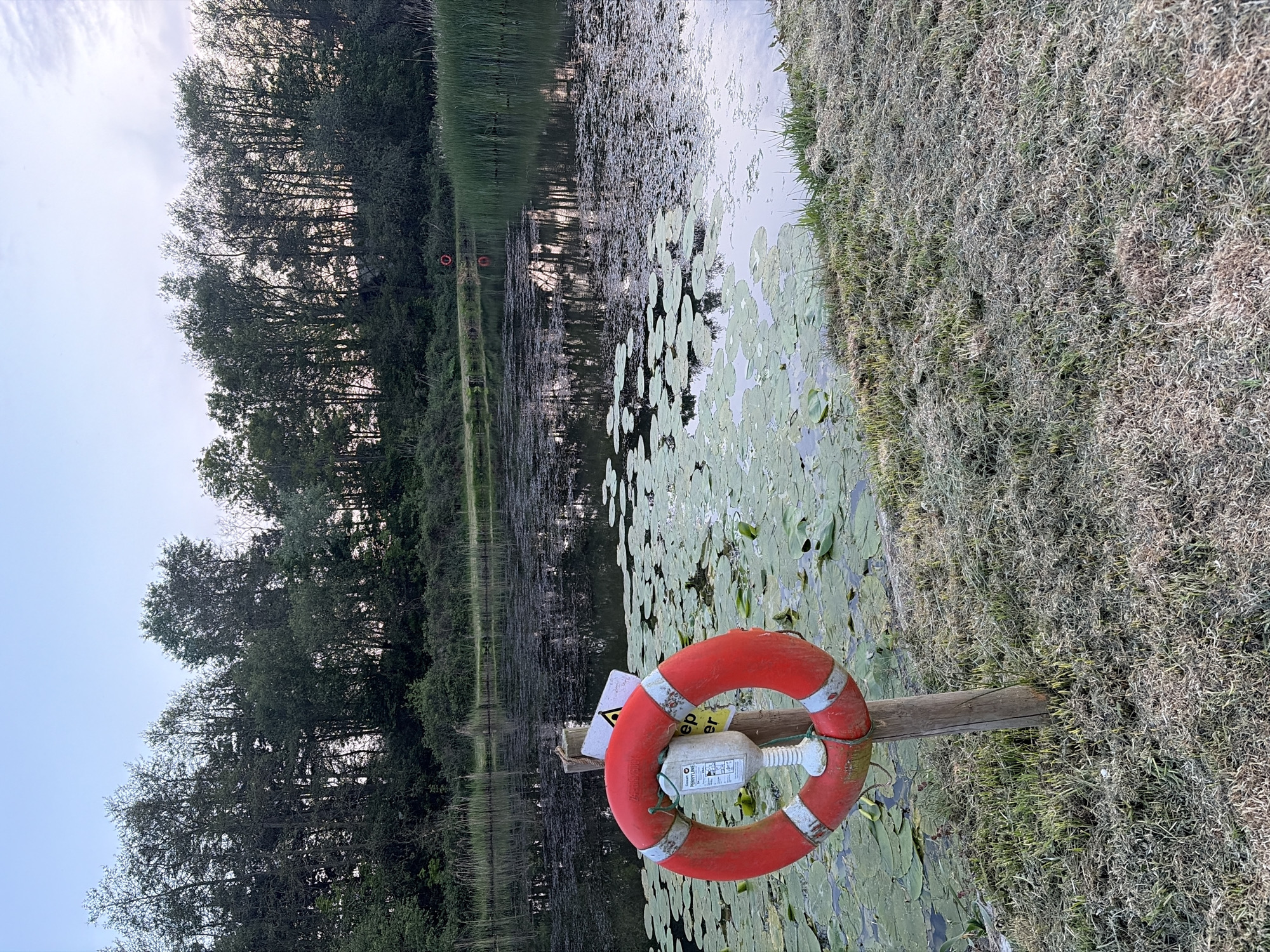 The image shows a body of water with lily pads, bordered by trees and grassy vegetation. A red life preserver hangs on a wooden post near the water's edge. The scene suggests a lakeside or pond environment. While the location is said to be East Suffolk, United Kingdom, there are no elements in the image to positively identify the exact location.