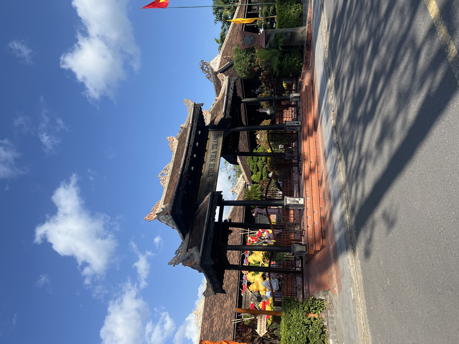 The image displays the grand, traditional Vietnamese-style entrance to a facility identified by a prominent sign as "VINH NHA TRANG" in the city of Nha Trang, Vietnam. The entrance structure consists of multiple dark-stained wooden pavilions with ornate, multi-tiered, dark brown tiled roofs featuring upward-curving eaves and decorative ridge tiles.
In the foreground, an asphalt road runs horizontally, bordered by a grey concrete sidewalk. Long, dark shadows, consistent with strong sunlight, are cast across the road and sidewalk. A five-step stairway, constructed from reddish-brown tiles, leads up to the main entrance area, which is partially secured by a black metal fence.
To the right of the entrance steps, a tall flagpole displays the red national flag of Vietnam with a yellow star. Adjacent to the steps on the right, a dark, sculptural monument is present amidst greenery. To the left of the steps, a well-maintained hedge of green bushes flanks the entrance, and a partially visible sign indicating "PHÒNG BÁN VÉ" (Ticket Office) and "BEVERAGE AREA" is visible.
Through the archway, various interior elements are discernible: additional traditional tiled roofs, extensive yellow and red festive decorations that include large, stylized figures, and abundant green foliage and potted plants. One individual in light-colored clothing is visible within the entrance area. Further inside on the right, a white statue and a yellow flag are visible.
The sky above is clear blue with scattered white cumulus clouds, indicating bright daylight conditions.