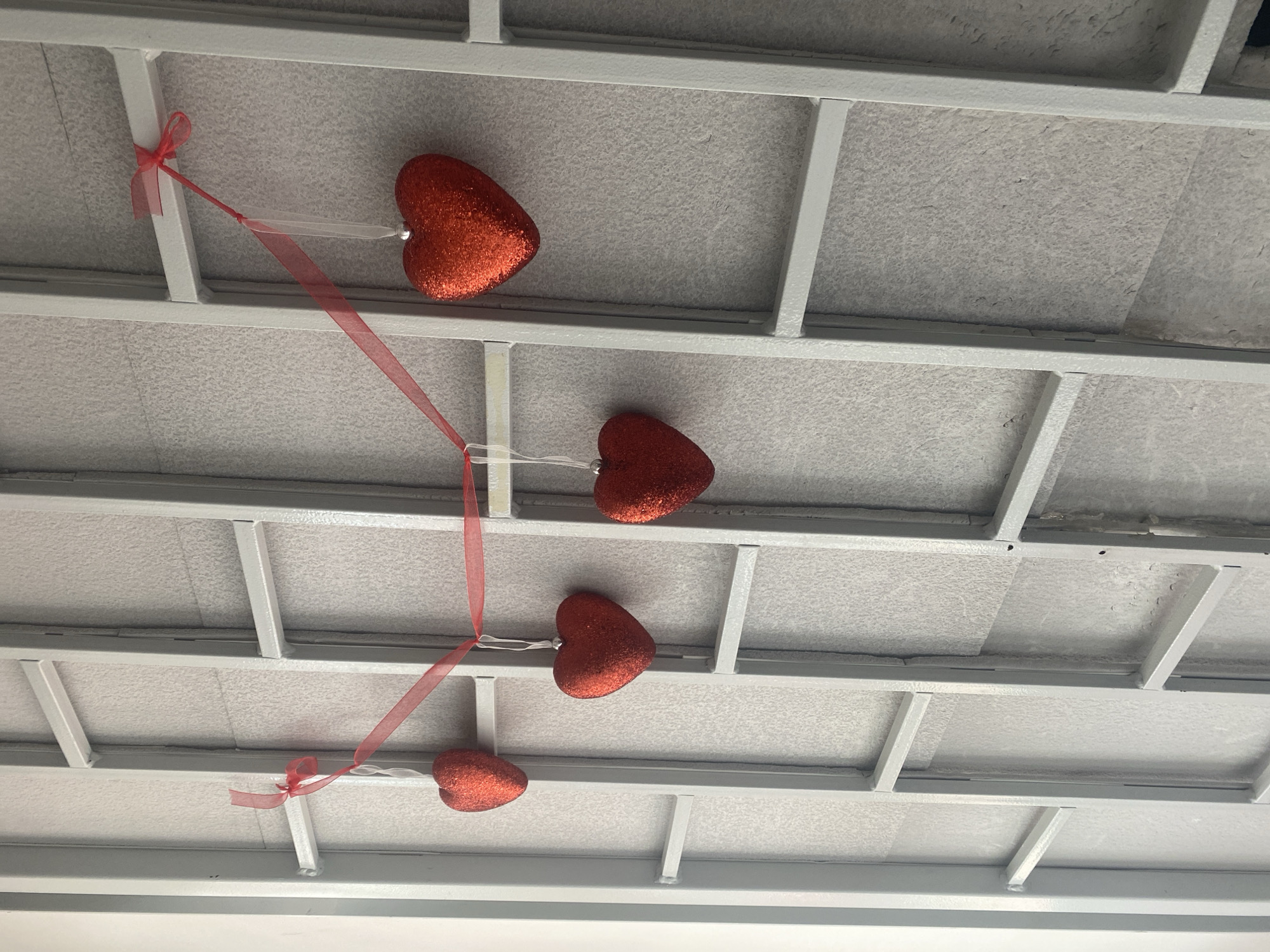 The image shows a ceiling made of white metal beams and textured ceiling tiles. Four red, glittery heart-shaped ornaments are hanging from the ceiling by thin ribbons. The top heart is attached to a white beam with a red ribbon tied in a bow. A long red ribbon extends down from the first heart, with two more hearts hanging from it at intervals. The bottom heart is attached to the ribbon with a small white string and a red bow. The scene appears to be indoors, possibly a room decorated for a celebration like Valentine's Day. The lighting is even, suggesting it might be daytime with artificial lighting. There are no people visible in the image. The location context is Novi Petrivtsi, Ukraine.
