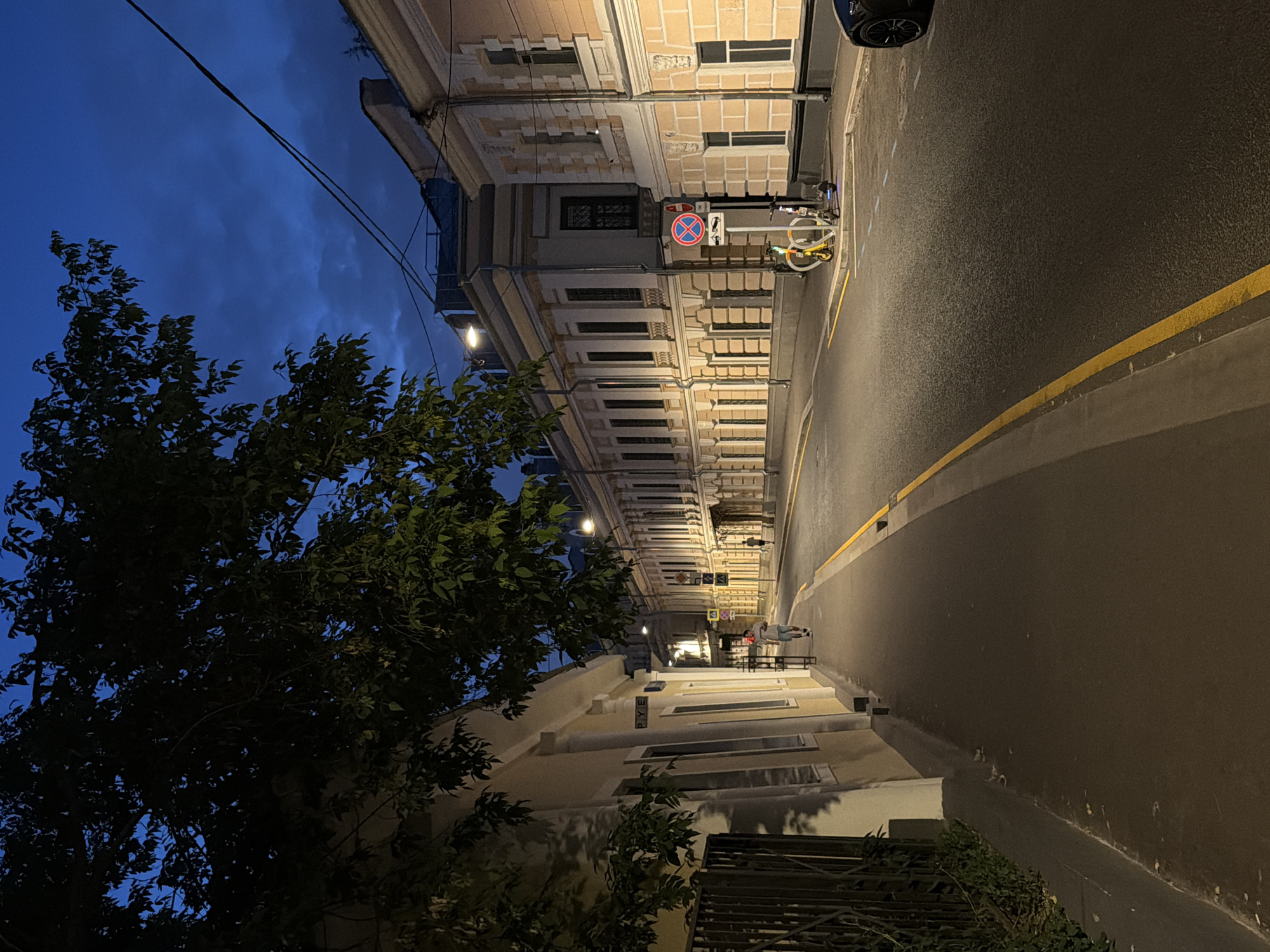 This image depicts a dimly lit street in Moscow, Russia, at dusk or early night. The sky is a deep blue, with some scattered clouds visible. The street is lined with old, ornate buildings, characterized by their light yellow facades, arched windows, and decorative cornices.
On the left side of the street, a sidewalk is visible, bordered by a dark metal fence and lush green foliage. A single person is walking away from the camera on this sidewalk. Further down the street, another person is seen walking.
The street itself is paved and illuminated by streetlights, casting a warm glow on the asphalt. On the right side of the street, parked bicycles and an electric scooter are visible, along with a traffic sign. A black car is partially visible on the far right edge of the frame. The overall atmosphere is quiet and serene, suggesting a typical evening in a historic urban setting.