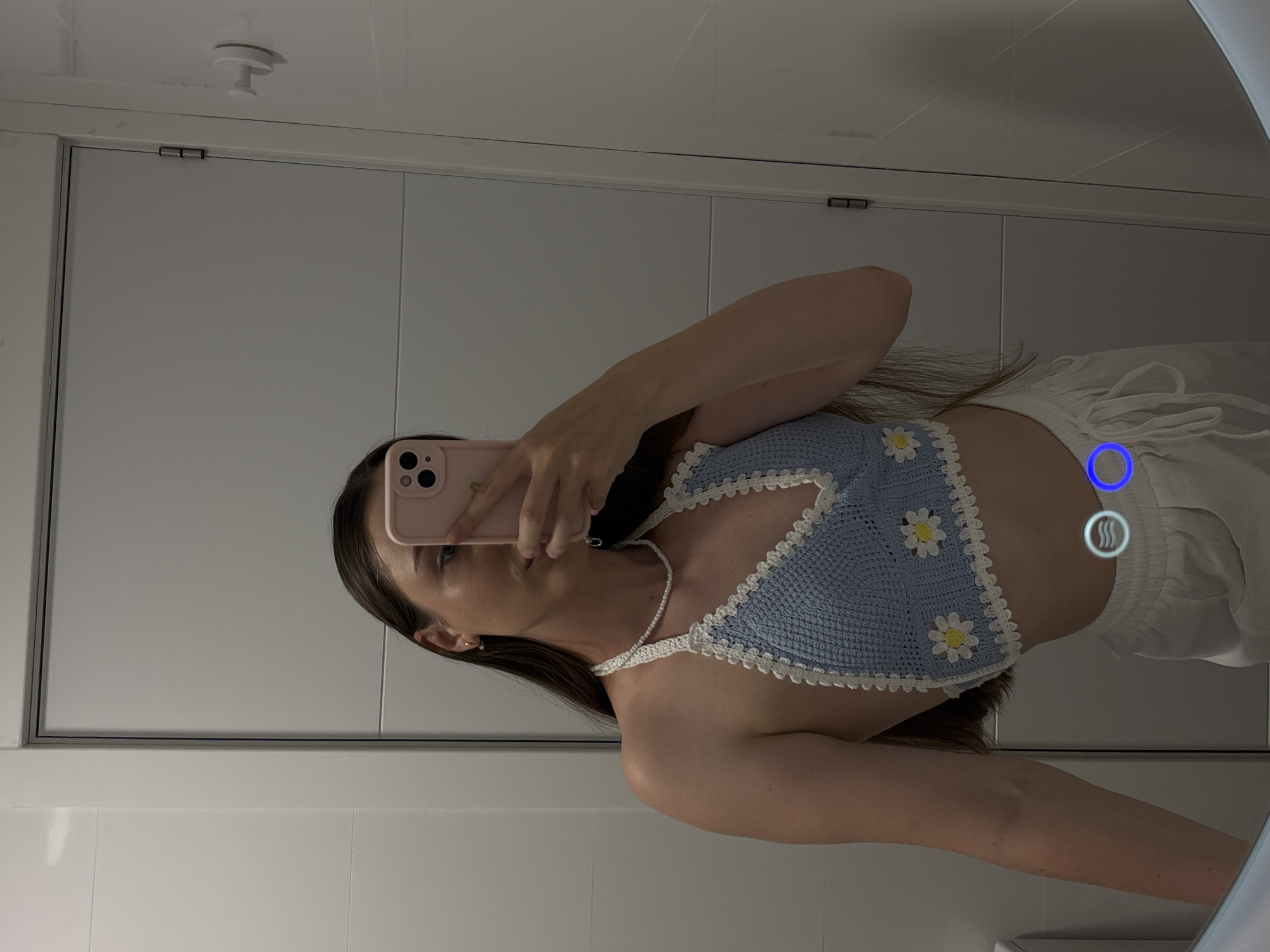 A young woman stands in front of a mirror, taking a selfie. She is wearing a light blue crochet halter top with white trim and embroidered daisies, and white drawstring pants. She has long, brown hair and is wearing a simple white beaded necklace and small gold hoop earrings. She is holding a pink iPhone to take the photo. The background appears to be a bathroom with white tiled walls and a door. There are two glowing blue circles on the lower part of the image, one with a wavy line inside and the other empty, which appear to be graphical overlays rather than part of the scene itself. The setting is a modern bathroom, likely in a private residence.