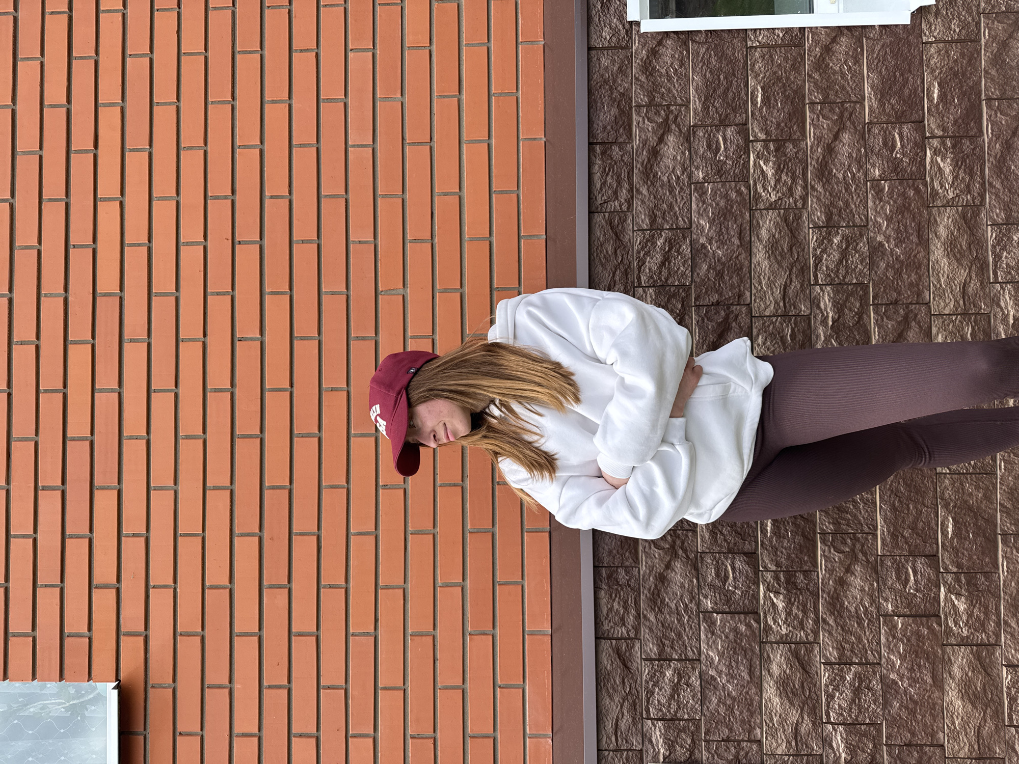 A sideways, medium shot depicts a woman in a burgundy baseball cap, a white sweatshirt, and brown leggings standing outdoors. She stands between a red brick wall and a wall of dark brown stone-like siding. The woman is facing towards the left of the image. She has her arms crossed in front of her. The location is Прикамский, Russia.