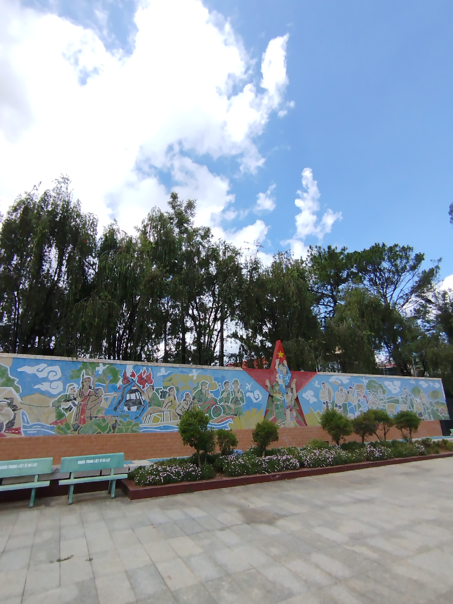The image captures a wide-angle view of a mural in Cam Ly - Đà Lạt, Vietnam, depicting historical scenes with figures and symbols relevant to Vietnamese history and culture. The mural stretches horizontally, with a central red star flanked by illustrations of people in various settings. The artwork is bordered by a low brick wall. In front of the mural, there is a landscaped area with small trees and flower beds. The foreground consists of a paved area and a green bench. Above the mural, trees partially obscure the sky, which is blue with scattered clouds.