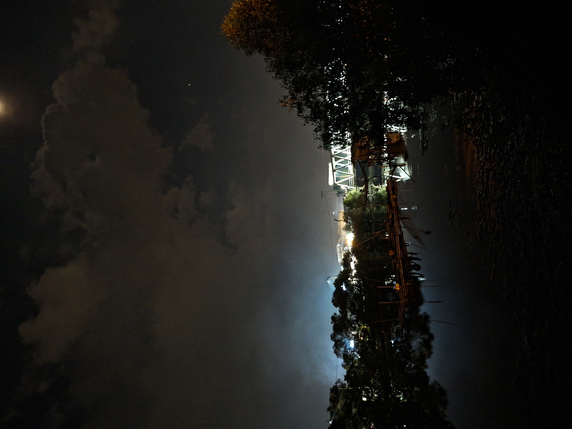 This nocturnal image, taken in Lhokseumawe, Indonesia, depicts a tranquil scene along a body of water, likely a river or canal, under a dark, cloudy sky. A bright, circular light source, possibly the moon, is visible in the upper left, partially obscured by clouds.
Along the right side, dense trees and foliage line the bank, silhouetted against the night. Numerous artificial lights illuminate various structures: a prominent, brightly lit white angular truss structure, possibly part of a bridge or large building, stands out in the upper right. Below this, other buildings or structures with lights are visible, some appearing to be constructed near the water's edge.
A long pier, also dotted with small lights, extends from the right bank into the dark, calm water. Several figures are discernible on the pier, appearing to be people, some holding long poles, suggesting they are fishing. The artificial lights from the bank and the pier cast soft reflections on the water's surface. The overall scene conveys a quiet nighttime atmosphere by the water. No text is visible in the image.
