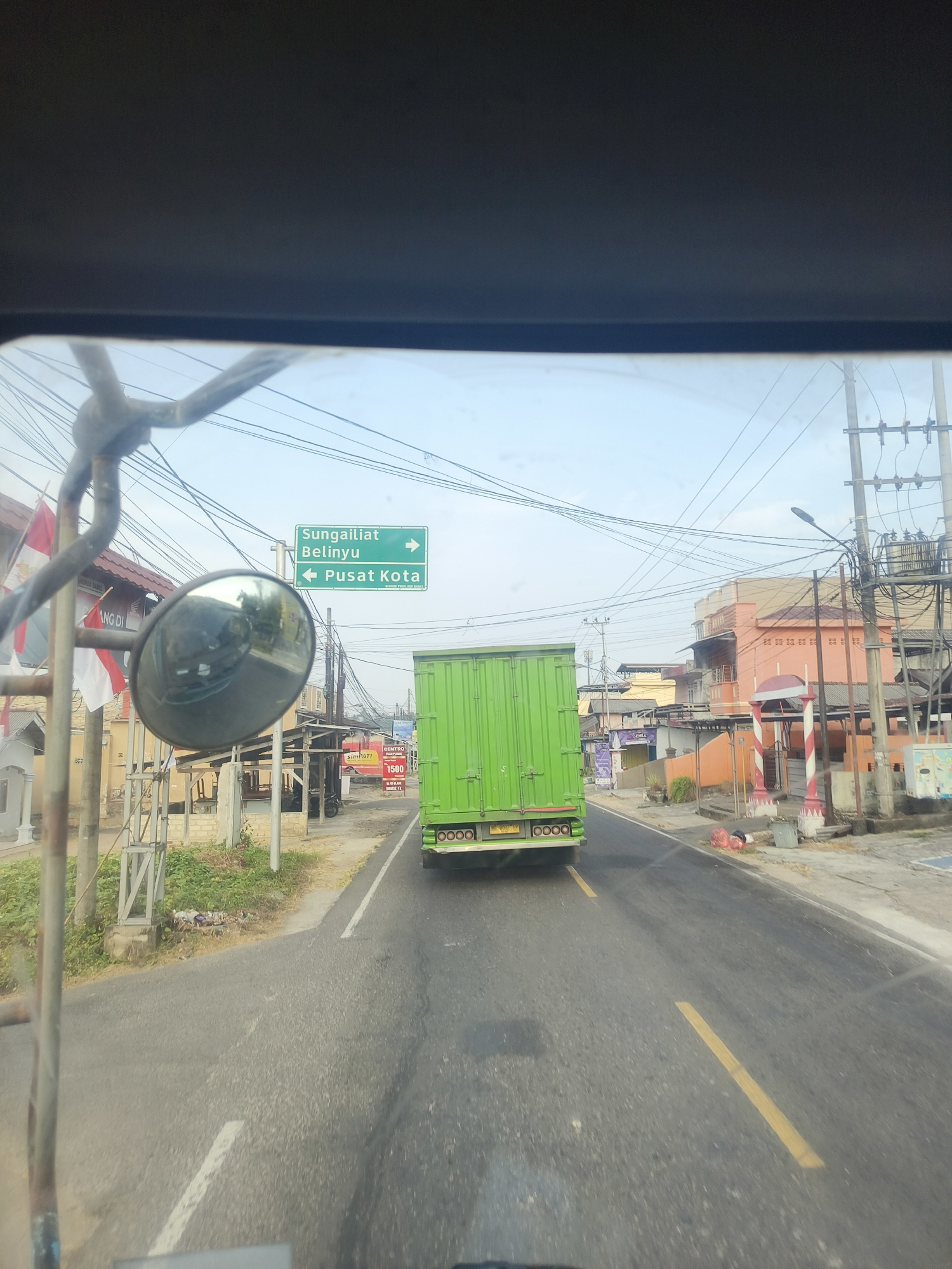 The image is taken from inside a vehicle, looking forward through the windshield. A large green truck is driving ahead on a road in what appears to be an urban or suburban area. There are buildings and power lines visible on the right side of the road. On the left side of the road, there are flags and a side mirror attached to a metal pole. A green directional sign is also visible on the left, indicating "Sungailiat" and "Belinyu" with arrows pointing to the right, and "Pusat Kota" with an arrow pointing to the left. The road has yellow lane markings. The sky is bright and appears to be daytime. The location is identified as Pangkalpinang, Indonesia based on the directional sign "Pusat Kota" which means "City Center" in Indonesian.