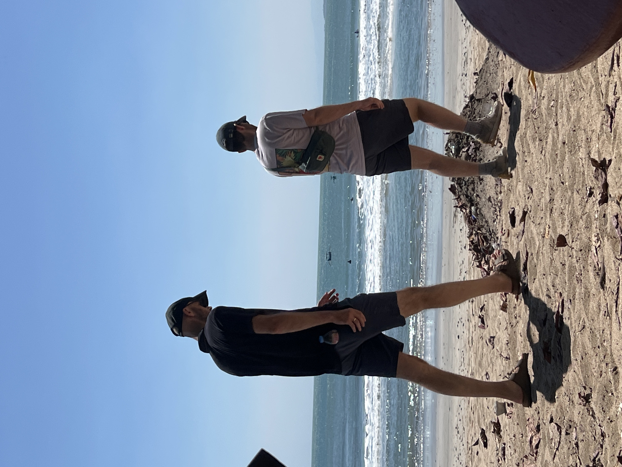 Two individuals, both wearing baseball caps and casual attire, are walking on a sandy beach in Kuta, Indonesia. The person on the left is wearing a black polo shirt and dark shorts, with a water bottle tucked into their pocket. The person on the right is wearing a light-colored t-shirt with a graphic print on the back and dark shorts, with a green fanny pack worn across their chest. They are both walking away from the camera towards the ocean, which is visible in the background with waves and several small boats. The beach is covered in sand and scattered debris, including dry leaves and what appears to be a pile of dirt or sand. The sky is clear and blue.