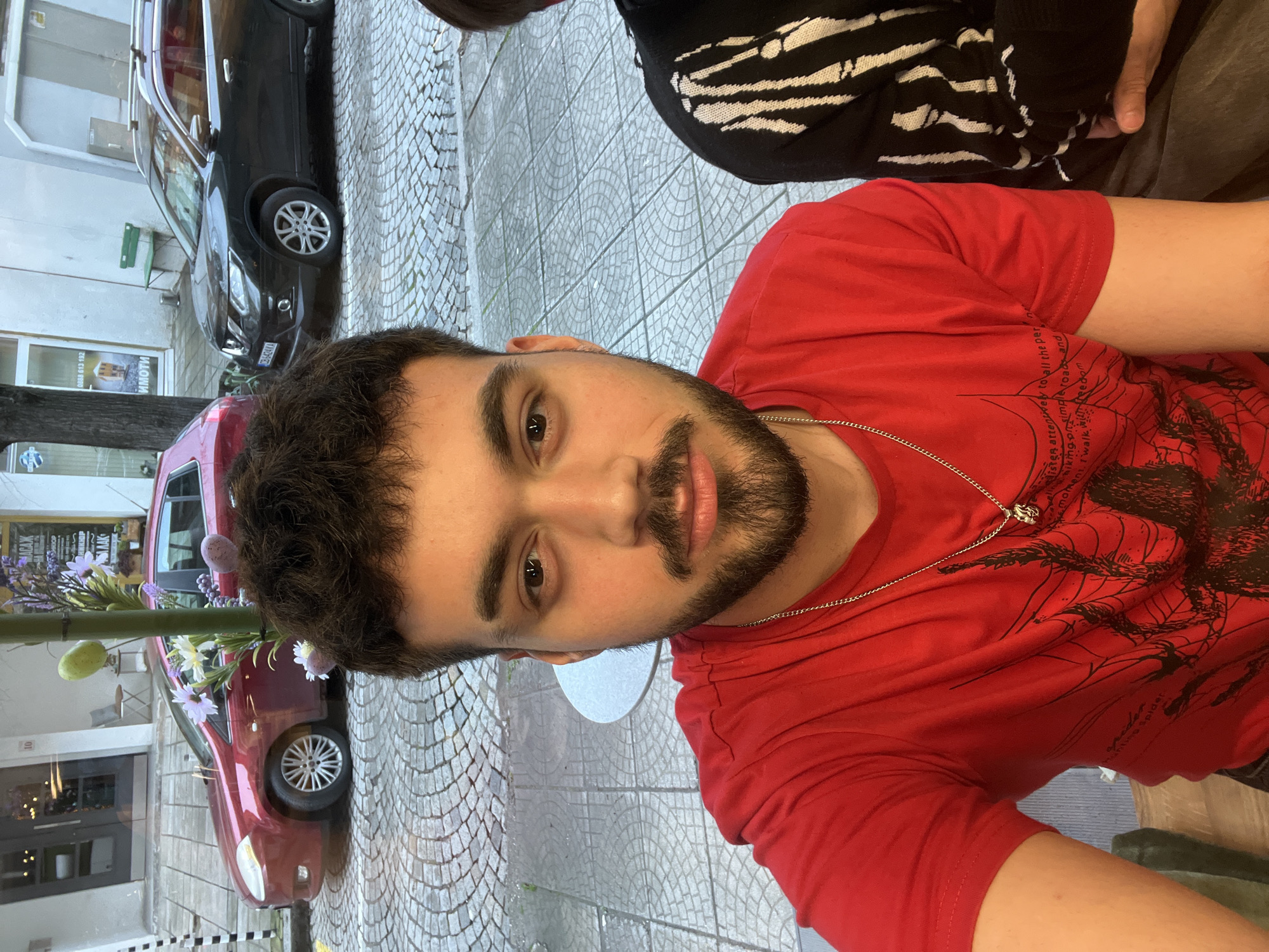 A man with dark hair and a beard is posing for a close-up photograph. He is wearing a red t-shirt with a graphic design and a silver chain necklace. The man is positioned outdoors, likely at a sidewalk café, as a round table edge is visible. The background features cars parked on a cobblestone street, with buildings visible in the distance. The image's OCR text suggests the location may be in Sofia, Bulgaria, based on the "Имоти" text and a local phone number. A flower decoration is placed on the left side of the image. Another person wearing a black and white striped garment is partially visible in the upper right corner. The street is paved with gray cobblestones.