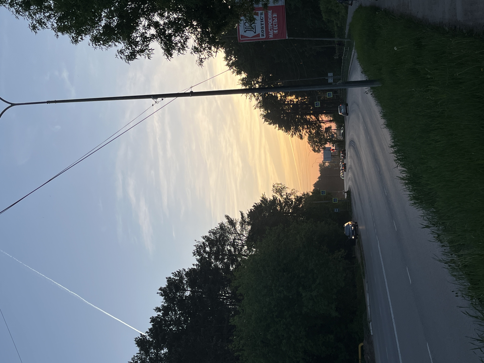 The image depicts a street scene in Chaikovsky, Russia, during a sunset. A road runs along the right side of the frame, with cars traveling in the distance. Lush green foliage borders the road. A utility pole extends vertically on the left, with wires stretching across the sky. The sky is a gradient of blue to orange, with light clouds scattered throughout. A contrail is visible across the blue portion of the sky. A Rostic's sign is visible among the trees on the right side of the frame. The trees appear to be densely packed.