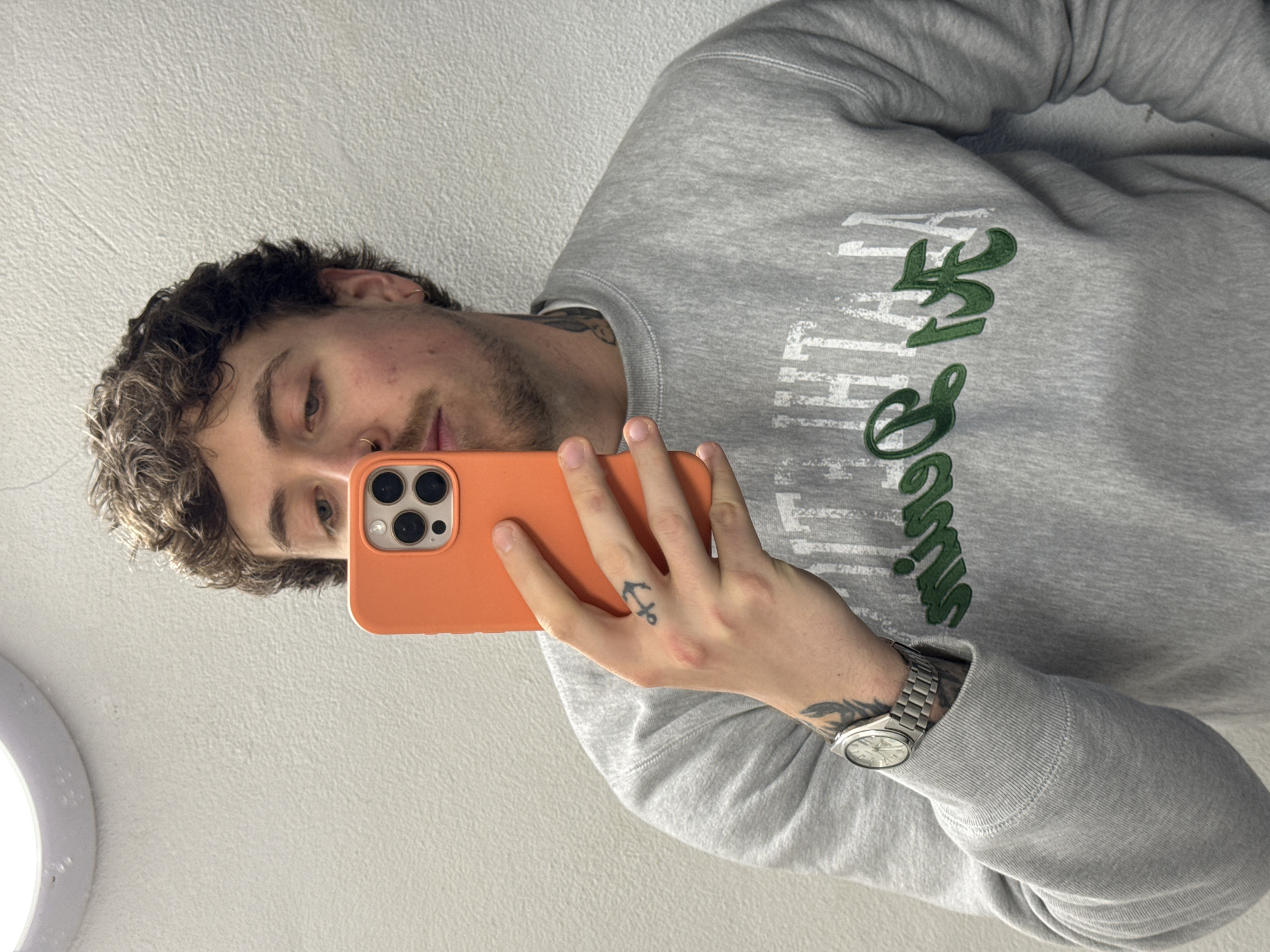 A young man is captured in a mirror selfie, positioned vertically against a light-colored, textured wall in an indoor setting, likely in London, United Kingdom. He is seen from the chest up, holding an orange smartphone with a prominent camera array in his right hand, reflecting his image.
The man has curly, dark brown hair, a light beard or stubble, and blue or green eyes. He wears a small hoop nose ring in his left nostril. His expression is neutral as he looks towards the phone screen.
He is wearing a grey sweatshirt adorned with distressed green and white text across the chest, which is partially obscured by the angle. Visible tattoos include a small black anchor on his right ring finger and a larger, darker tattoo on his left forearm, partially covered by a silver-colored watch. Another tattoo is visible on his neck, peeking out from beneath the sweatshirt collar.
In the bottom left corner of the reflection, a bright circular light fixture, possibly a ring light, is visible. The overall lighting suggests an indoor environment.