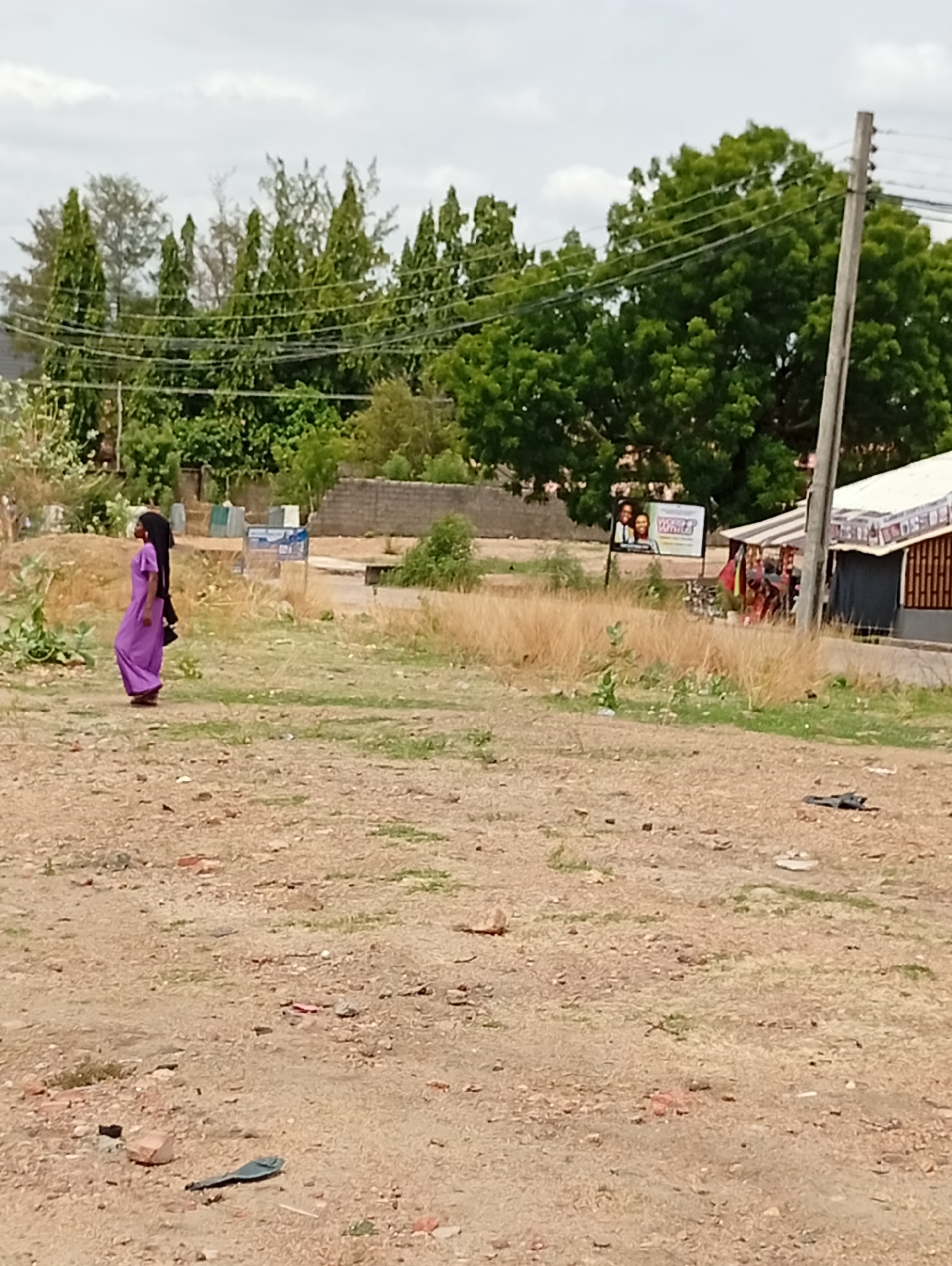 A medium, outdoor shot depicts a sandy clearing in Adankolo, Nigeria. In the mid-left of the frame, a woman in a purple dress with a black head covering is walking left-to-right. The ground is mostly bare dirt with scattered debris. In the background, there are various trees and shrubs, a concrete block wall, and power lines overhead. To the right, a small roadside shop is visible with a partially obscured sign next to it. A utility pole stands prominently in the right background. The sky is overcast.