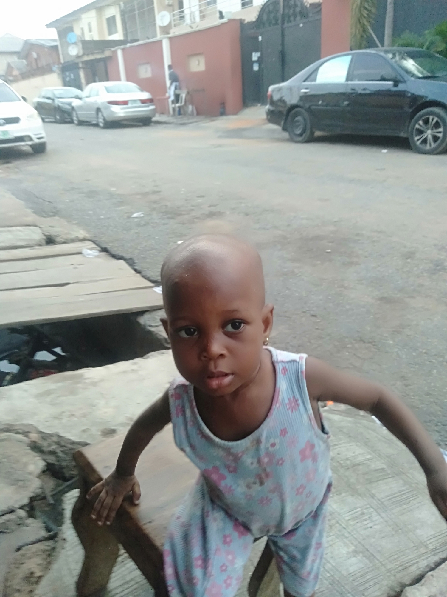 A young child wearing blue and pink floral patterned pajamas stands on what appears to be a wooden bench or low table outdoors. The background shows a street scene with several cars parked along the side, buildings, and a pedestrian. The location is the city of Shangisha, Nigeria. The child is the main subject of the image, looking directly at the camera. The setting suggests a residential area.