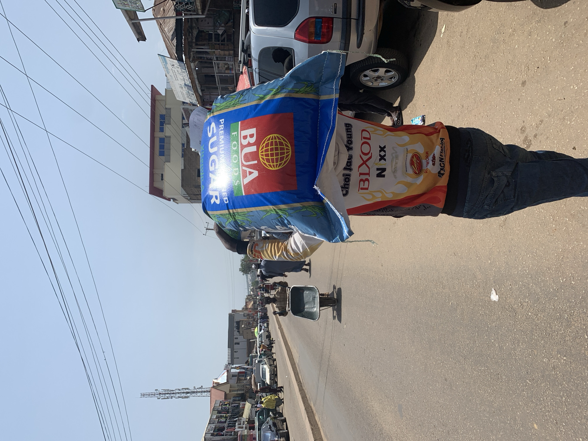 In an outdoor, daytime scene on a paved road, a man carries a large, blue bag of BUA Premium Sugar on his head. The bag is emblazoned with the BUA Foods logo, featuring a red circle with a yellow globe inside. The man wears a colorful shirt with graphics and dark denim jeans. He walks down the street, which is lined with buildings and stalls on the left, and vehicles, including cars and a silver SUV, on the right. Other pedestrians are visible in the background, as are utility poles and a cell tower. A man with a wheelbarrow is also seen in the mid-ground. The overall impression is that of a busy market or commercial area in the city of Jos, Nigeria, under a clear sky.