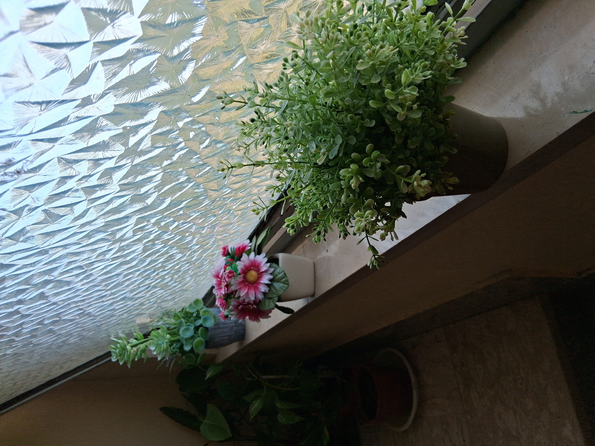 The image shows a close-up view of several artificial plants on a windowsill in Colorno, Italy. The windowsill is made of a light-colored, possibly marble, material and is positioned below a textured glass window. In the foreground, a dense, green artificial plant in a dark pot is visible on the right side of the frame. To the left and slightly behind it, a smaller pot holds a cluster of artificial pink and white flowers with a yellow center. Further to the left, another artificial plant with succulent-like leaves in a grey pot is present. On the far left, the dark foliage of a real plant in a brown pot is partially visible. The background consists of the patterned glass window, which refracts the light coming from outside, creating a textured and somewhat abstract visual effect. The overall scene suggests an indoor setting, likely a home or a public building in Colorno, where decorative plants are used to enhance the environment. No people are visible in the image.